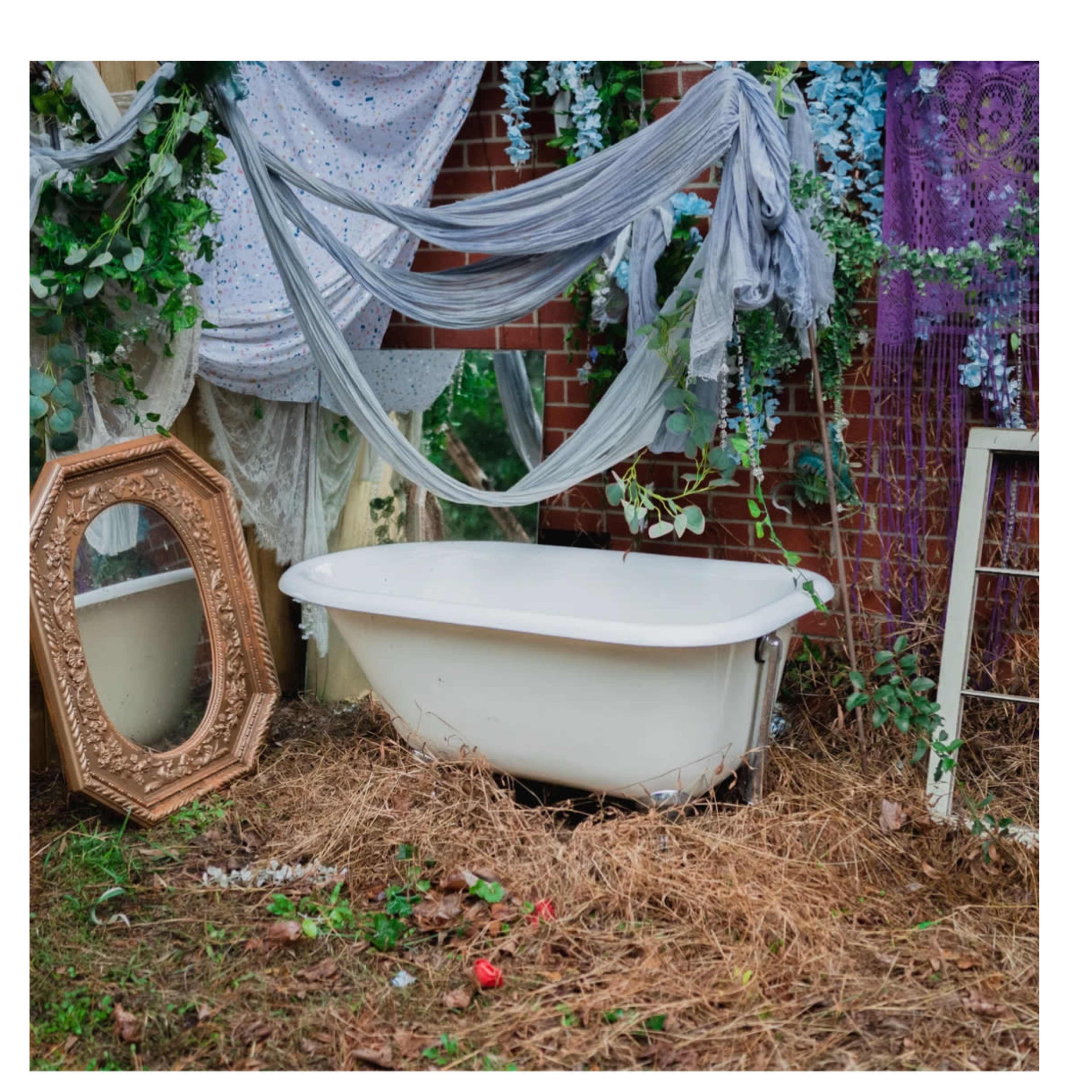 A vintage bathtub is situated in a lush garden setting adorned with fabric drapes and a large mirror nearby.