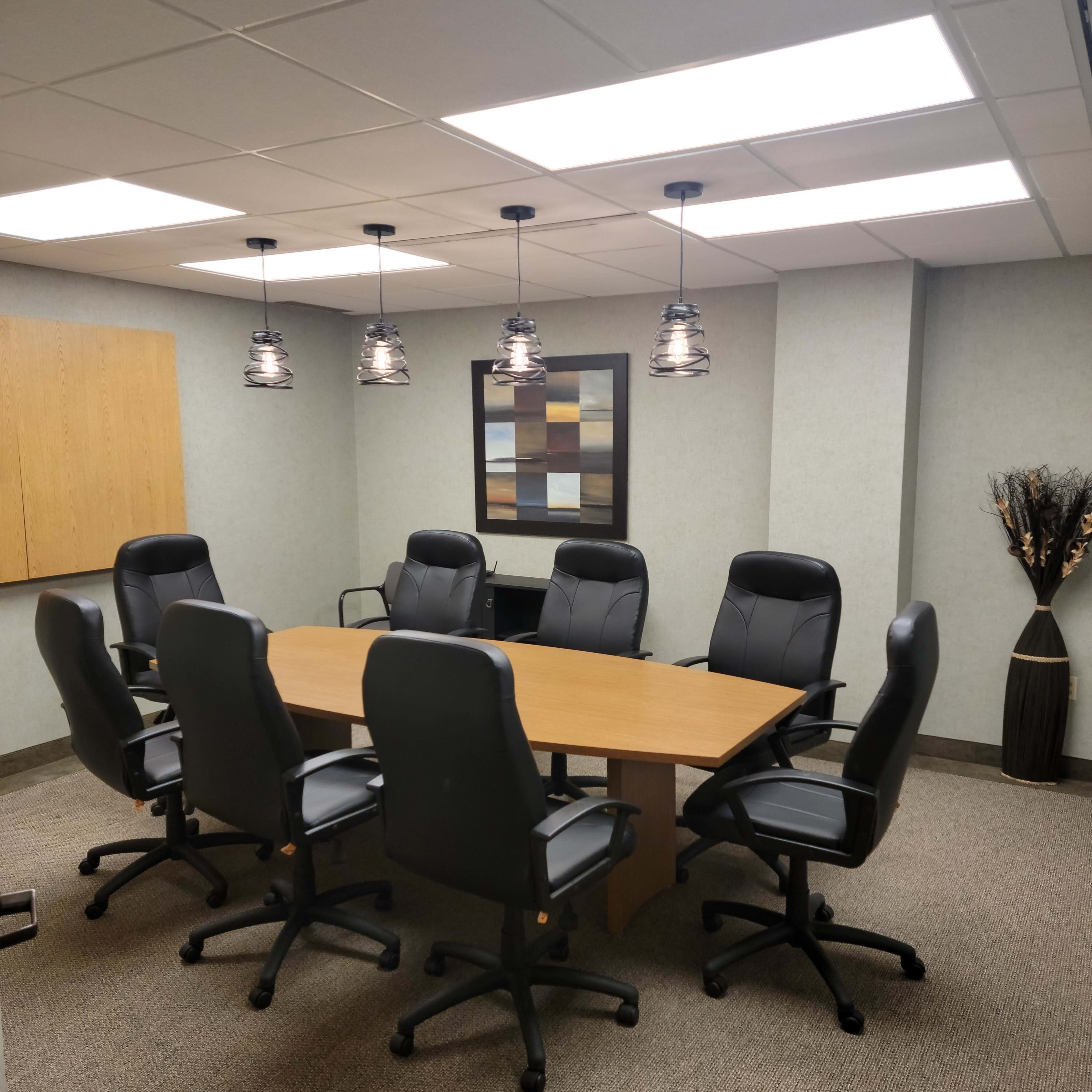A modern conference room features a large wooden table surrounded by six black office chairs and decorative lighting.