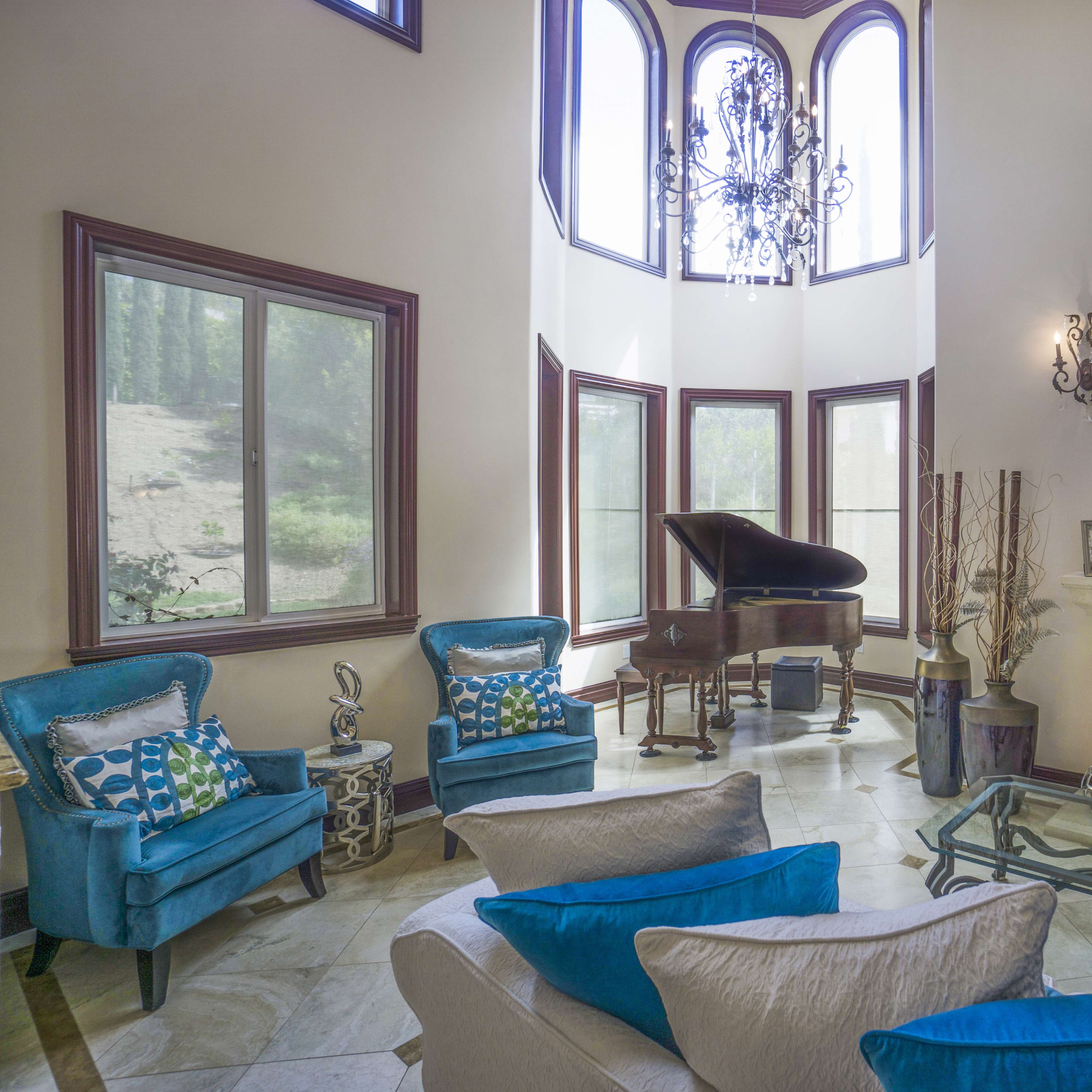 A spacious living room features a grand piano near large windows, with two blue chairs and a glass coffee table in front of a fireplace.