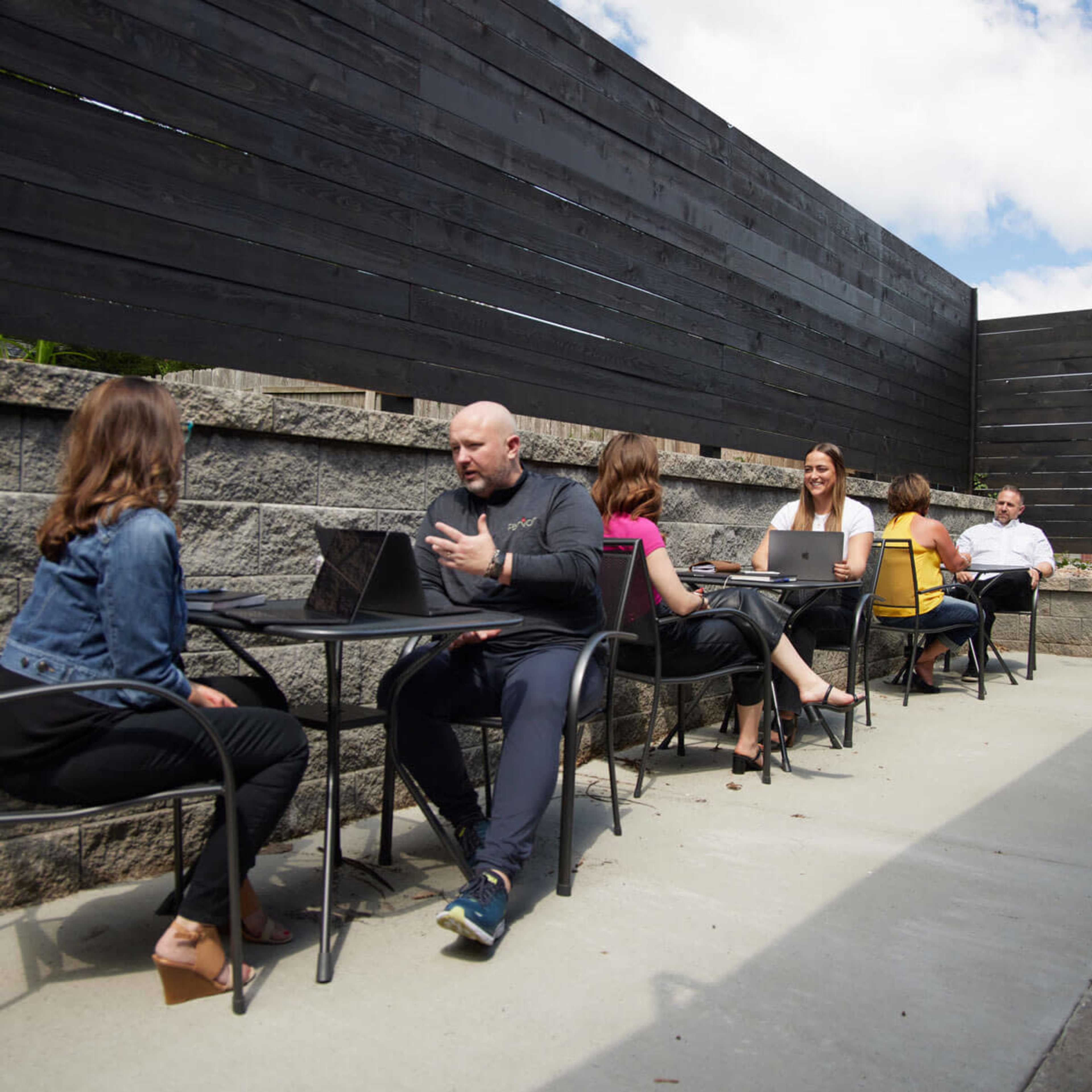 A group of six people are sitting at outdoor tables with laptops, engaged in conversation in a patio area.