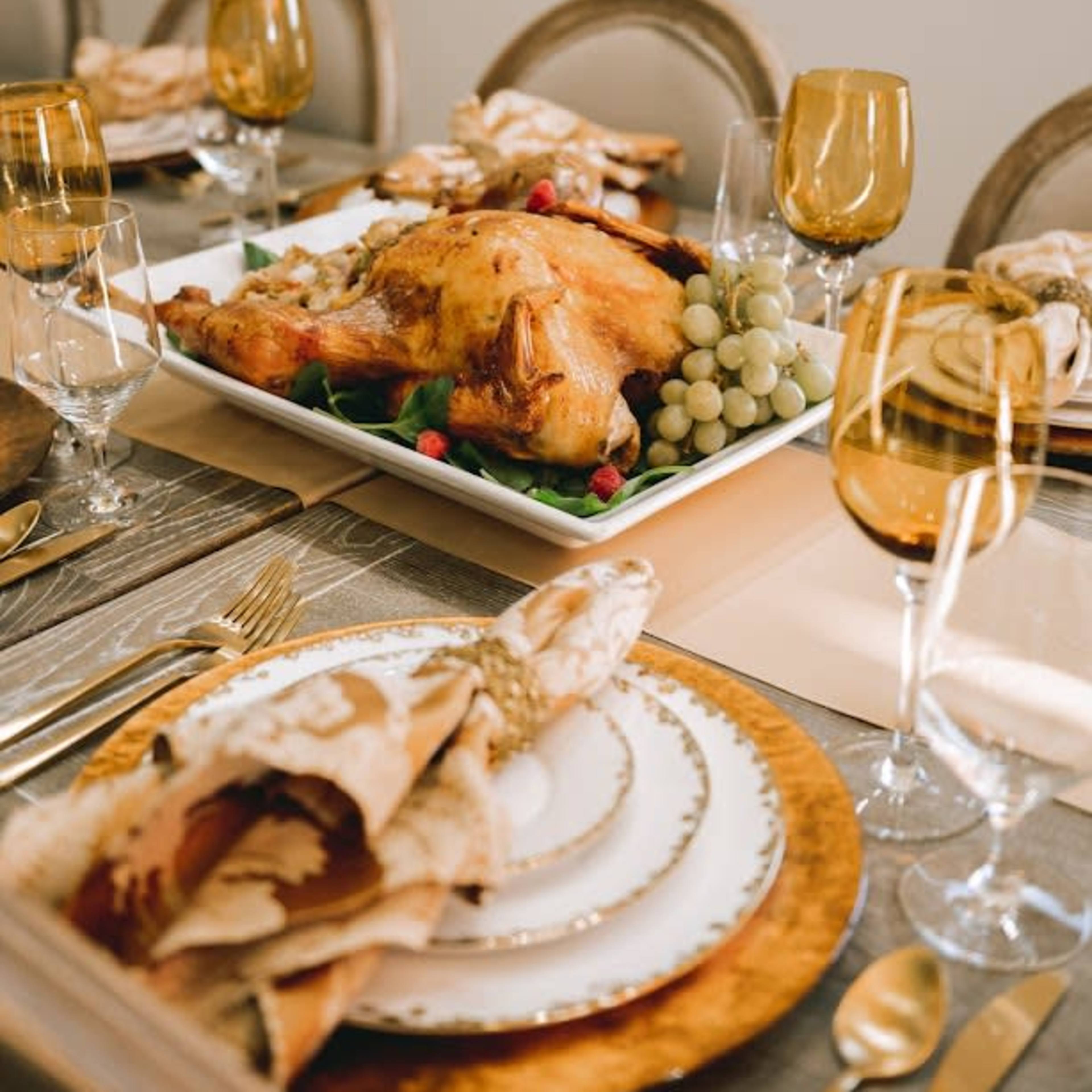 A beautifully arranged dining table features a roasted turkey on a platter surrounded by grapes and garnishes, with elegant tableware and golden glasses.