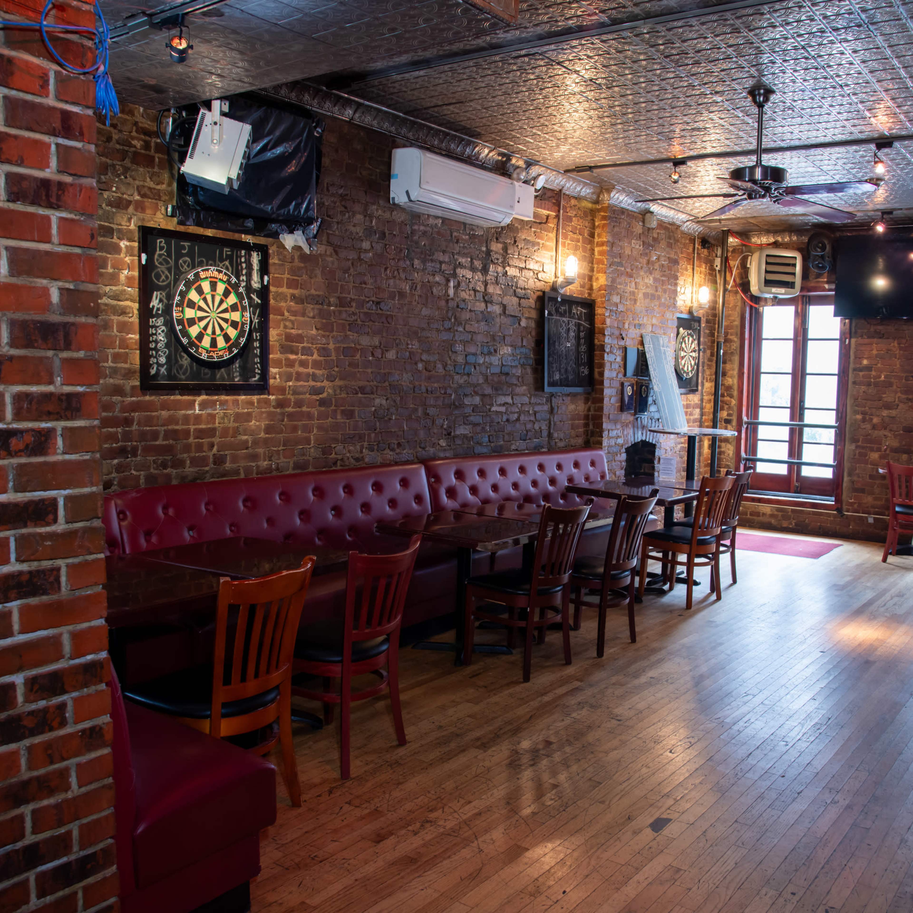 An empty bar interior with exposed brick walls, wooden tables, and a dartboard on the wall.