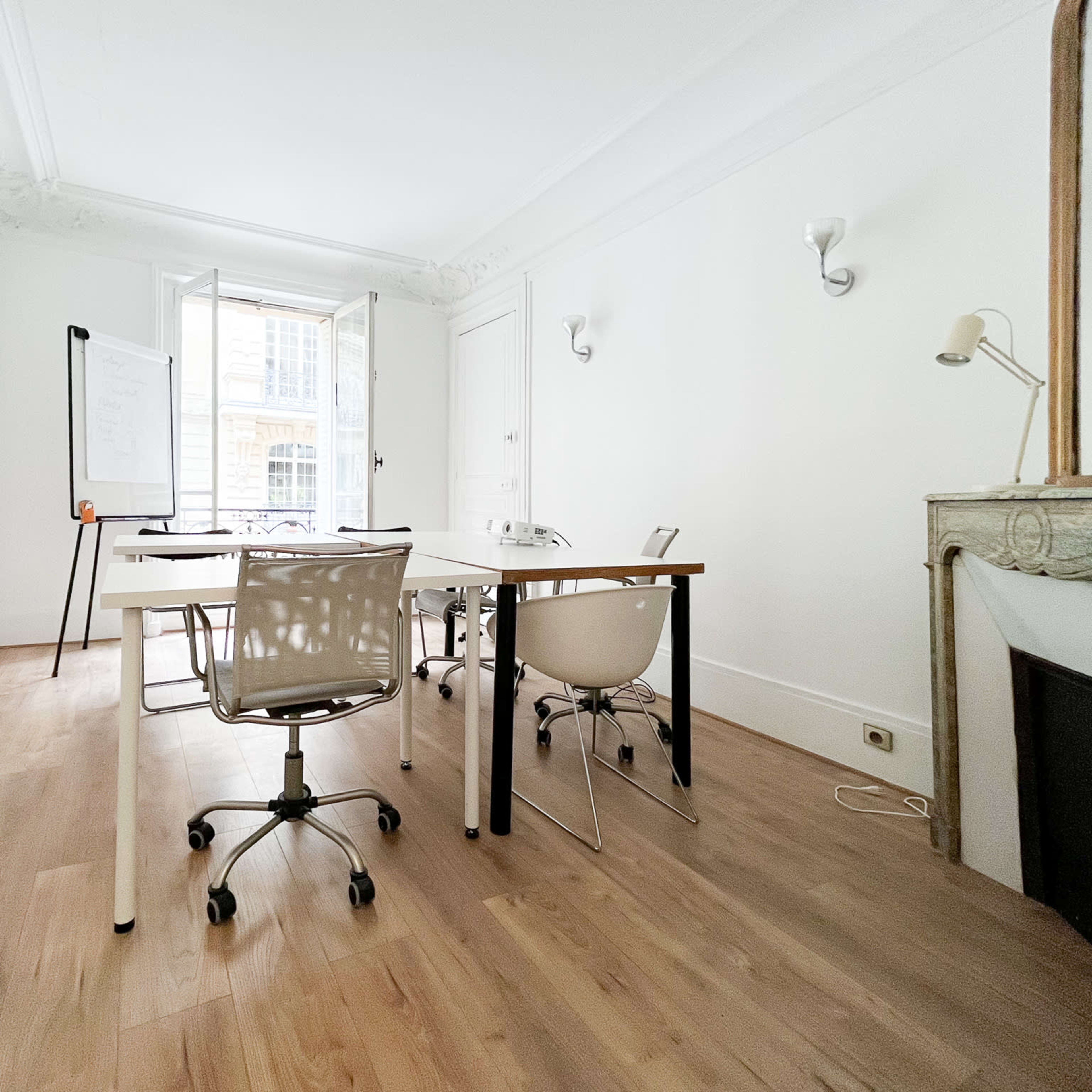 A bright, minimalist office space with a large window, a whiteboard on a stand, and two desks with chairs arranged around them.