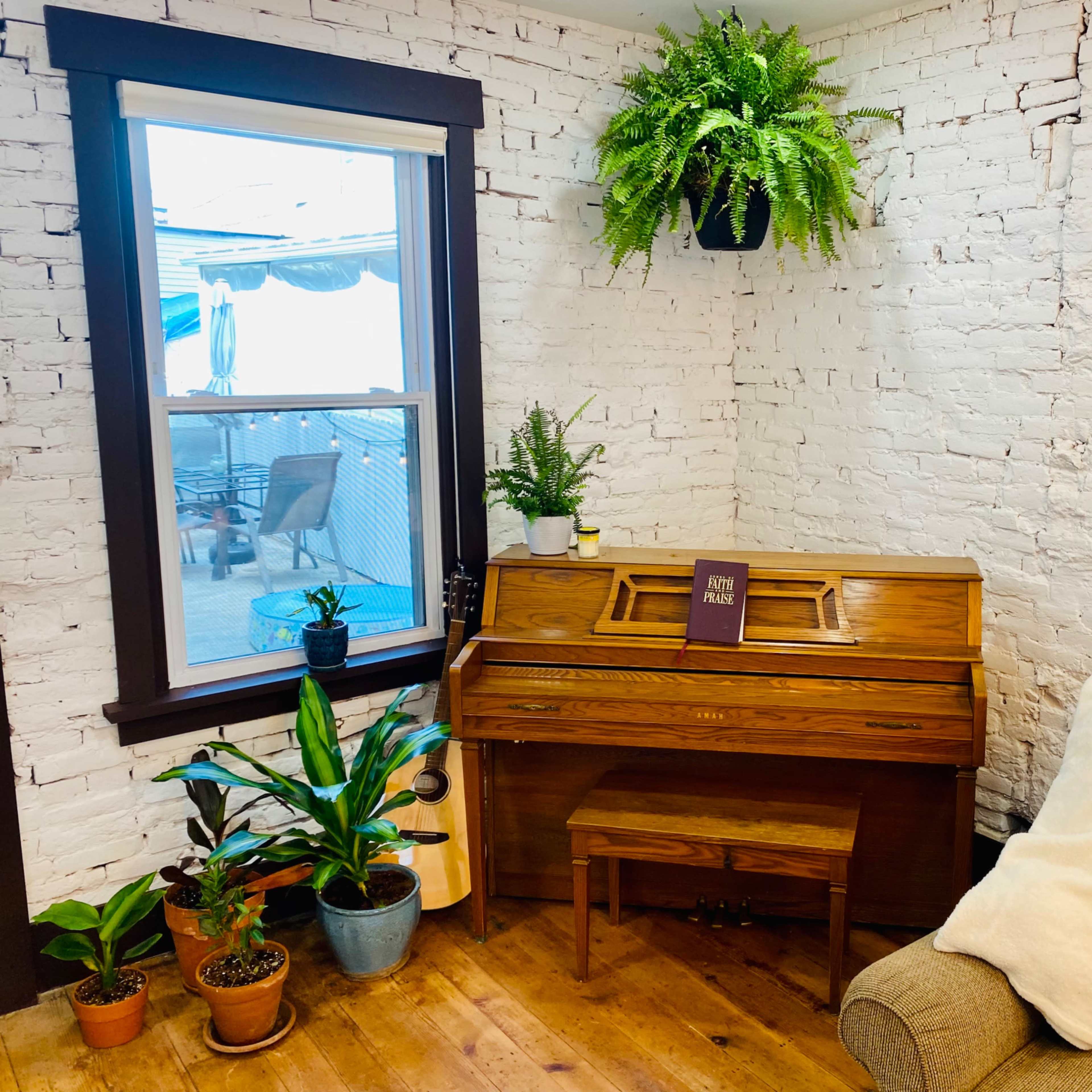 A wooden piano is positioned in a bright corner with potted plants and a decorative throw blanket on a tan sofa.