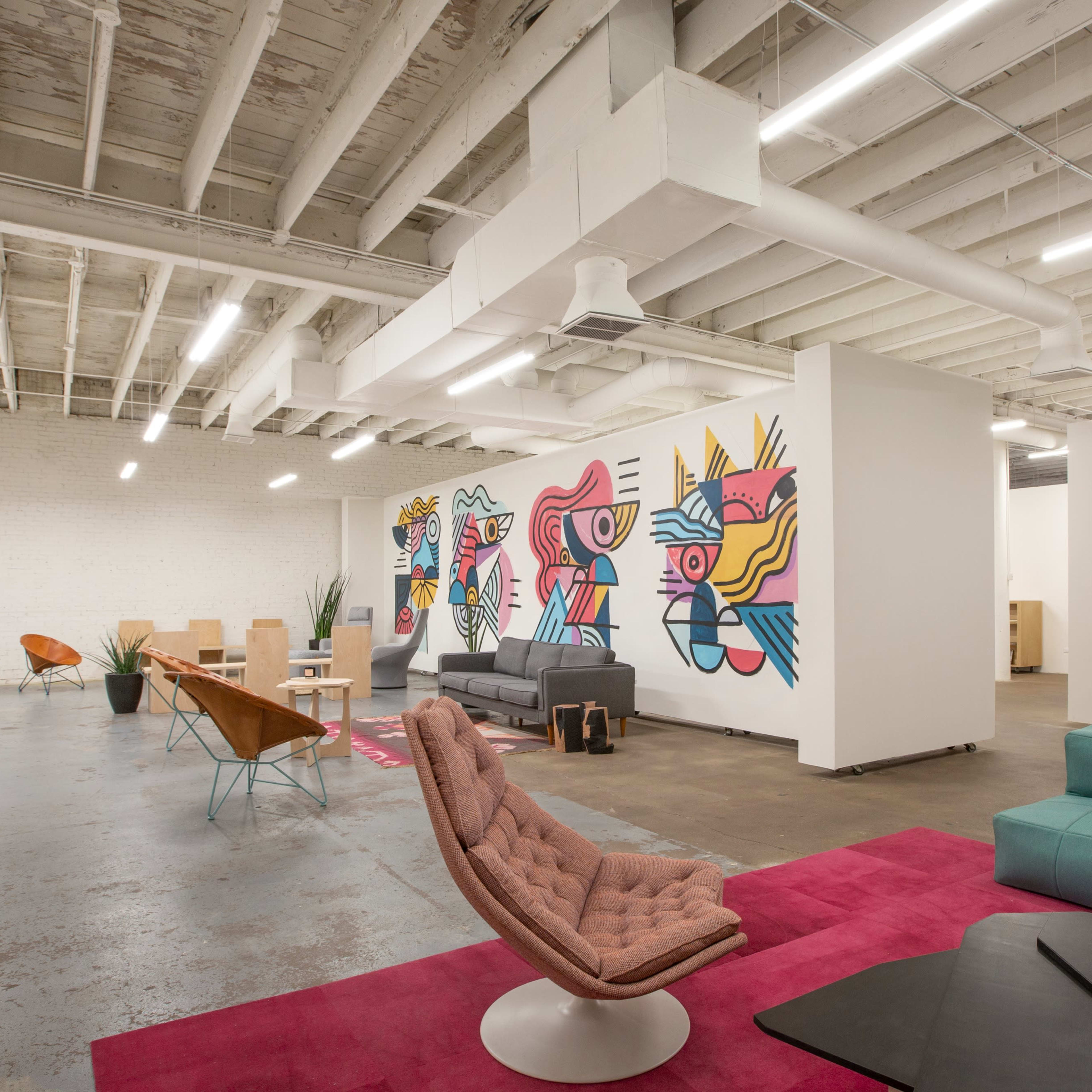 A modern office space featuring colorful wall art, various seating arrangements, and a combination of open areas and workstations.