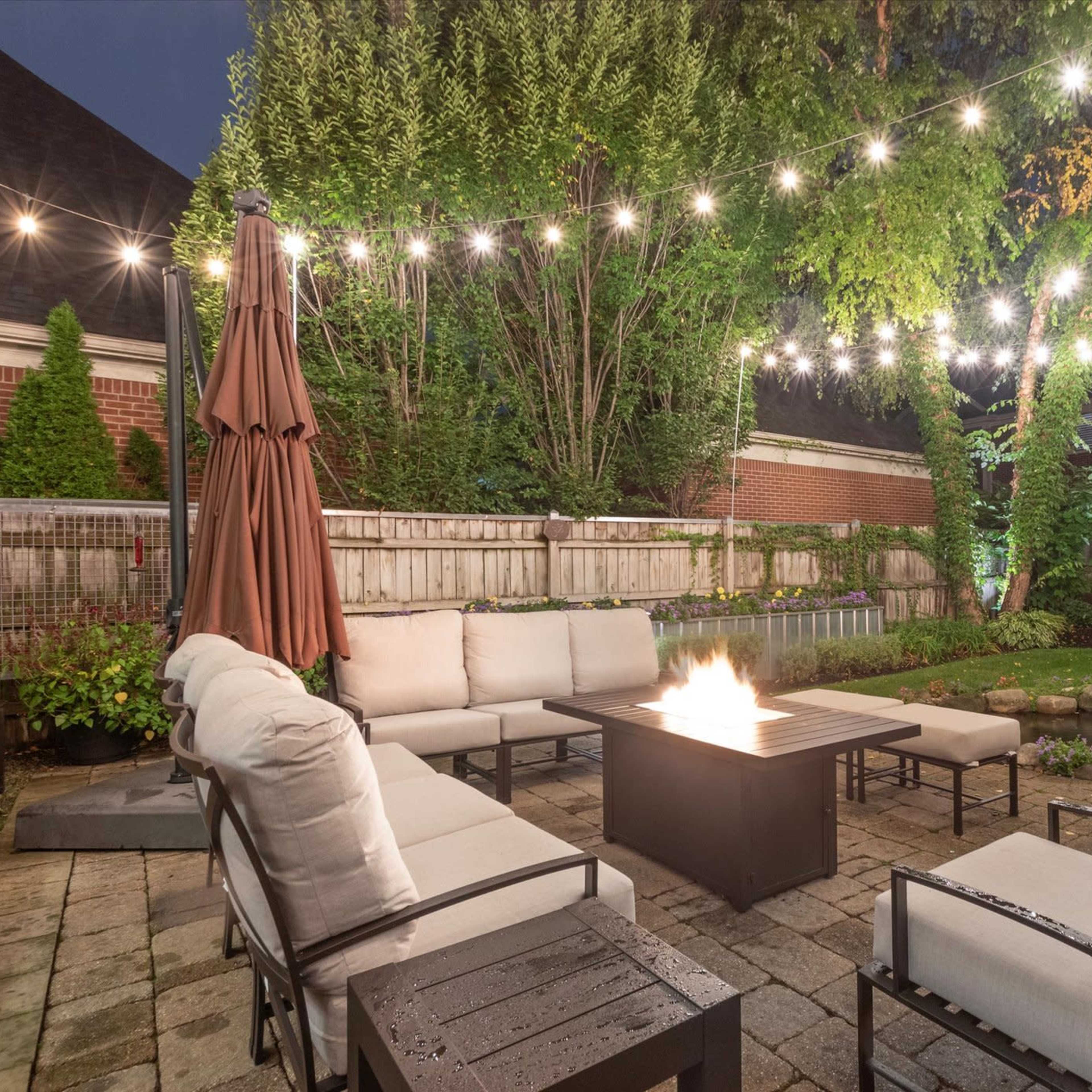 A patio area features a fire pit surrounded by seating, with string lights illuminating the space at dusk.
