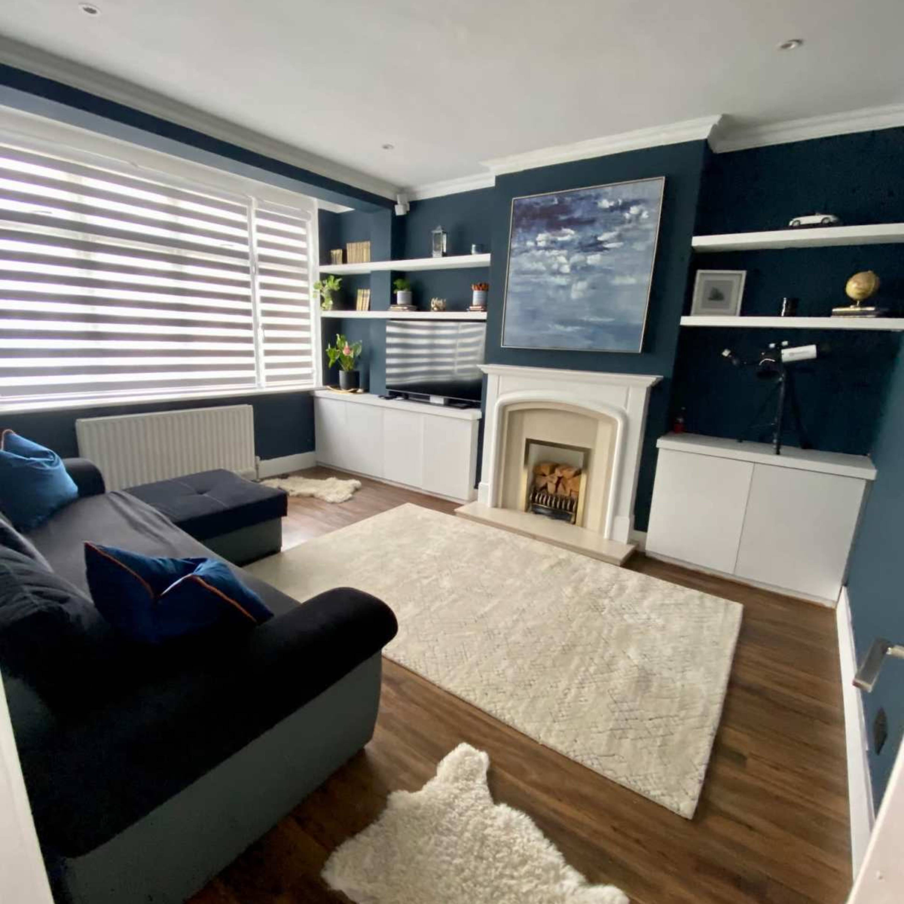 A modern living room with dark blue walls, a white fireplace, a large window with horizontal blinds, and a dark sofa facing a media center.