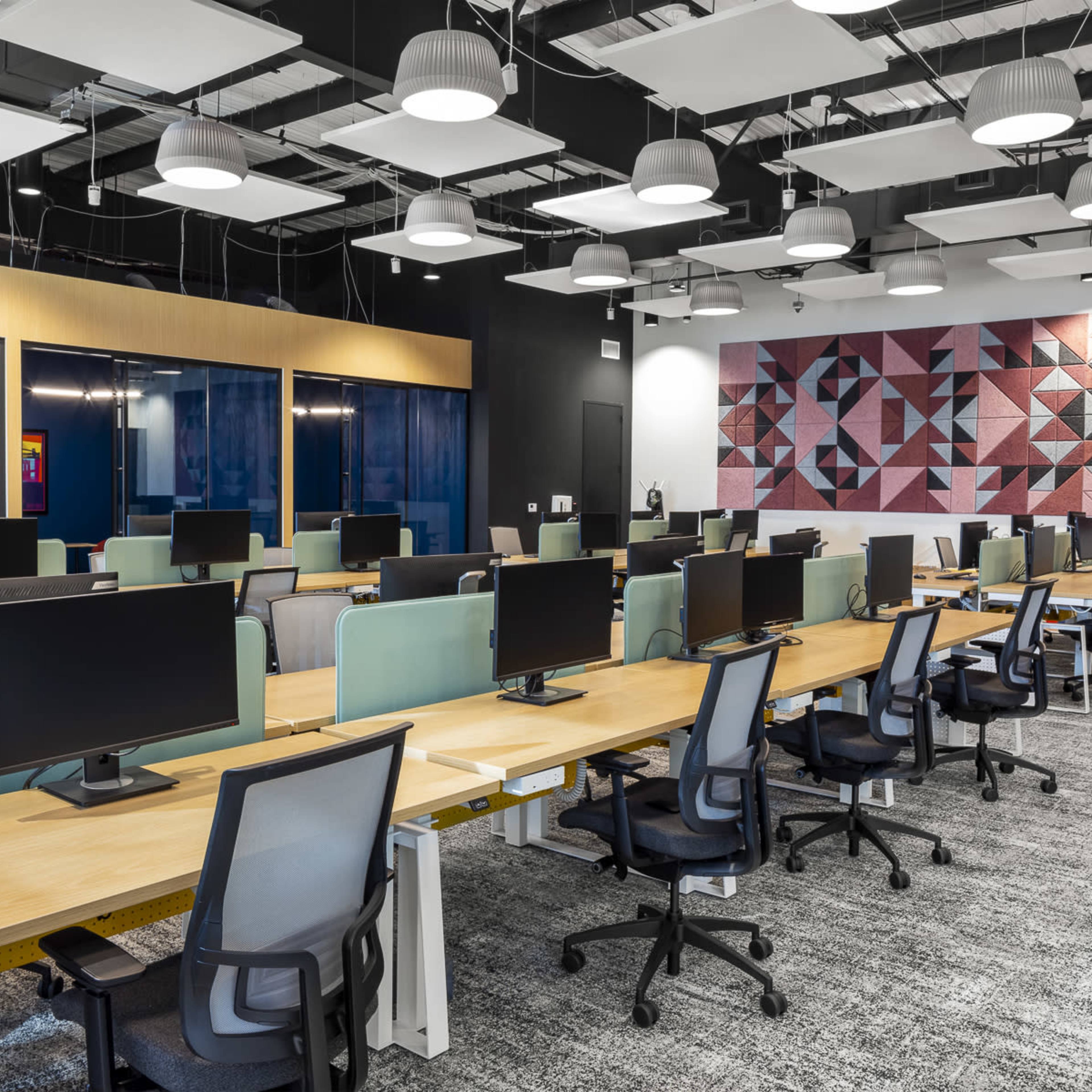 A modern office space with multiple workstations equipped with computers and ergonomic chairs, arranged in a grid pattern against a backdrop of colorful wall art.