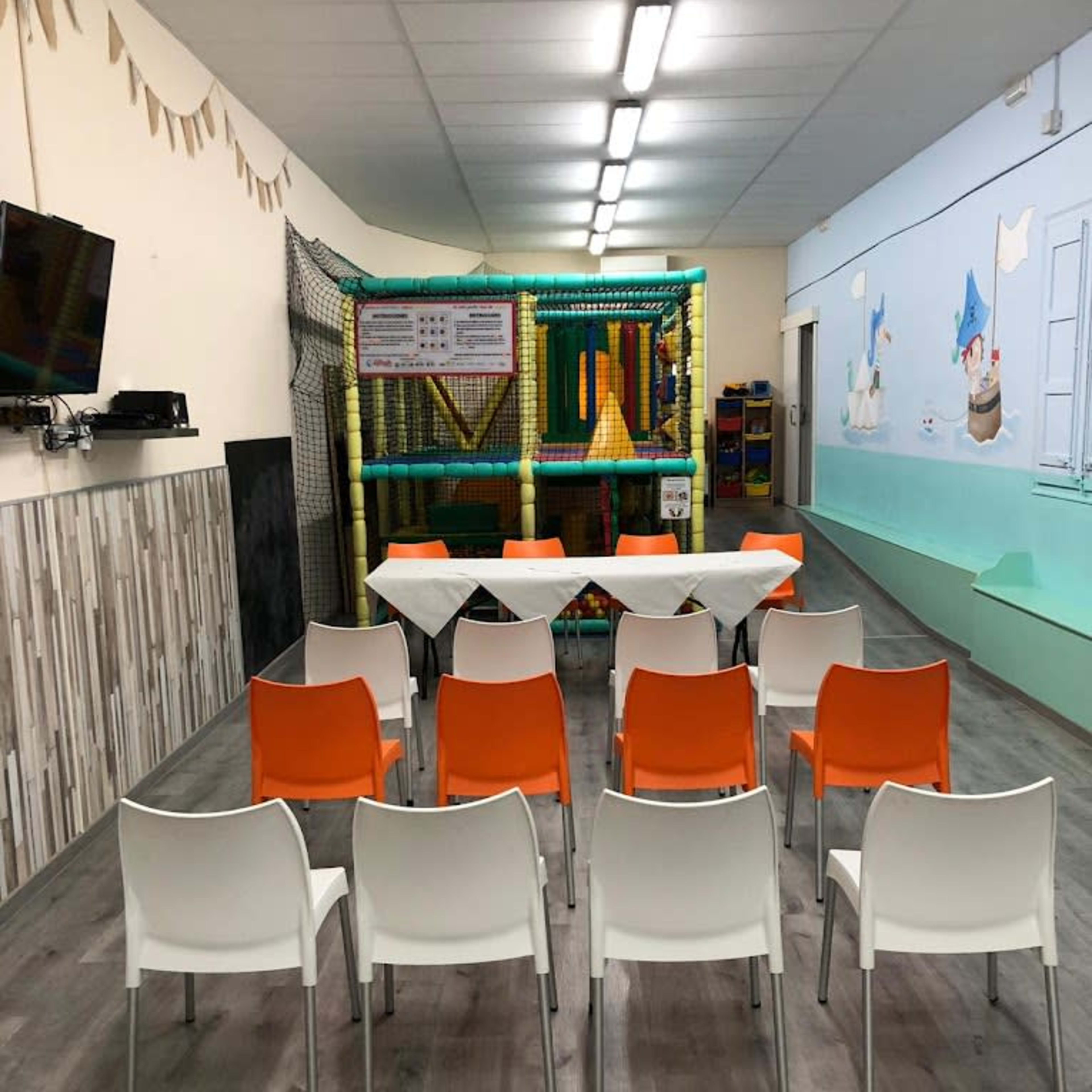 A spacious room with a play area in the back, arranged with a row of white and orange chairs facing a table.