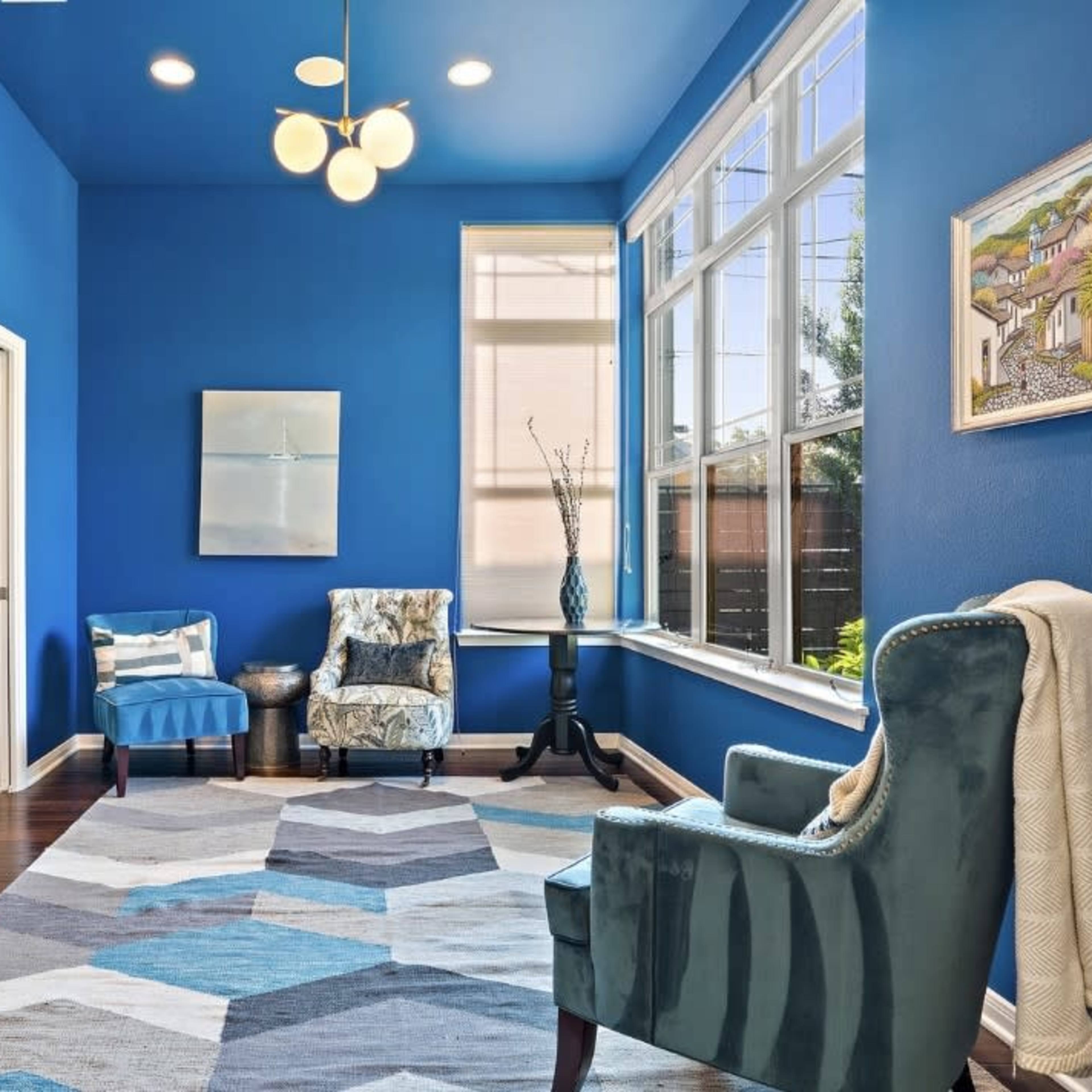 A brightly lit room features blue walls, a geometric area rug, two upholstered chairs, and a ladder against a shelving unit.