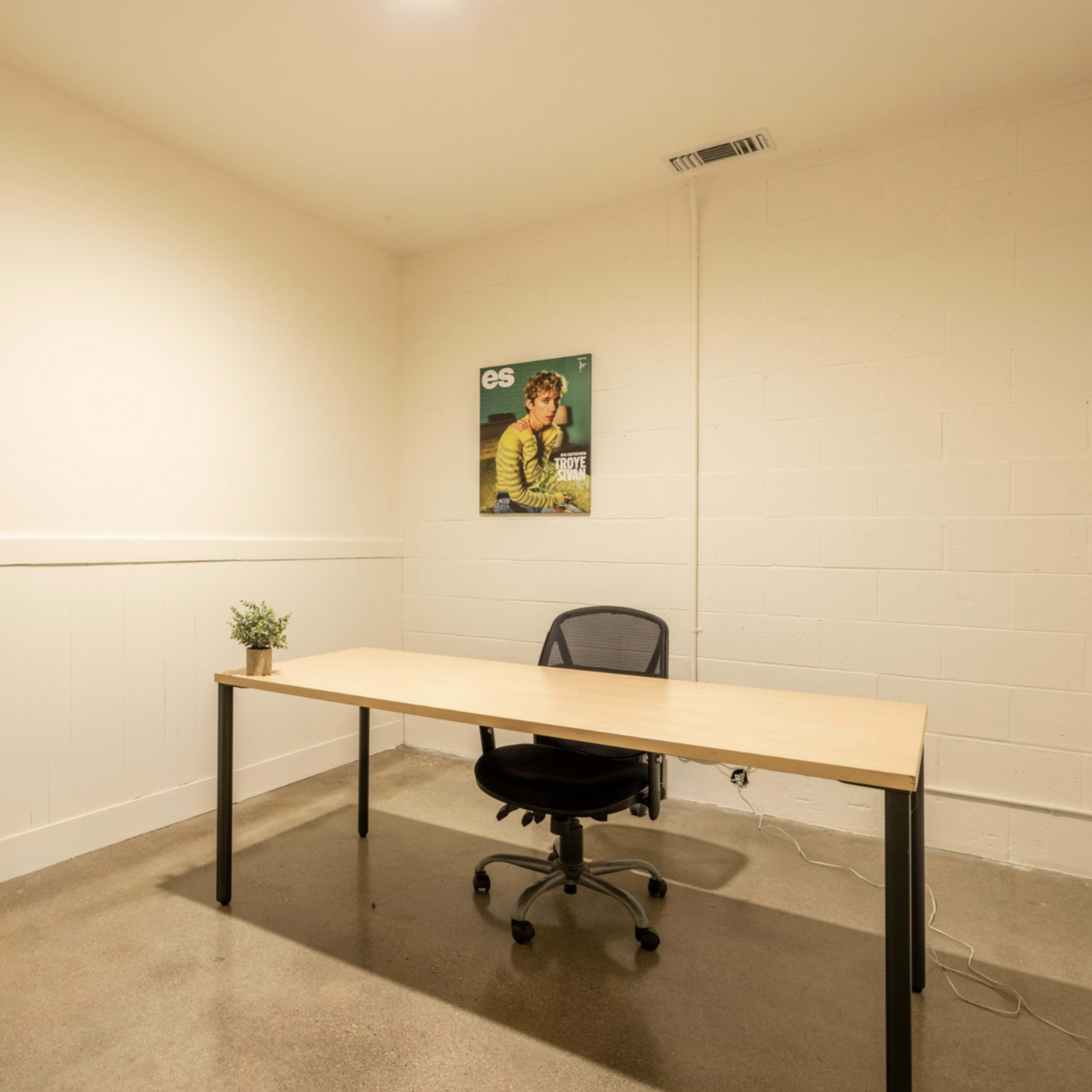 An empty office space features a wooden desk with a swivel chair and a small plant, alongside a poster on the wall.