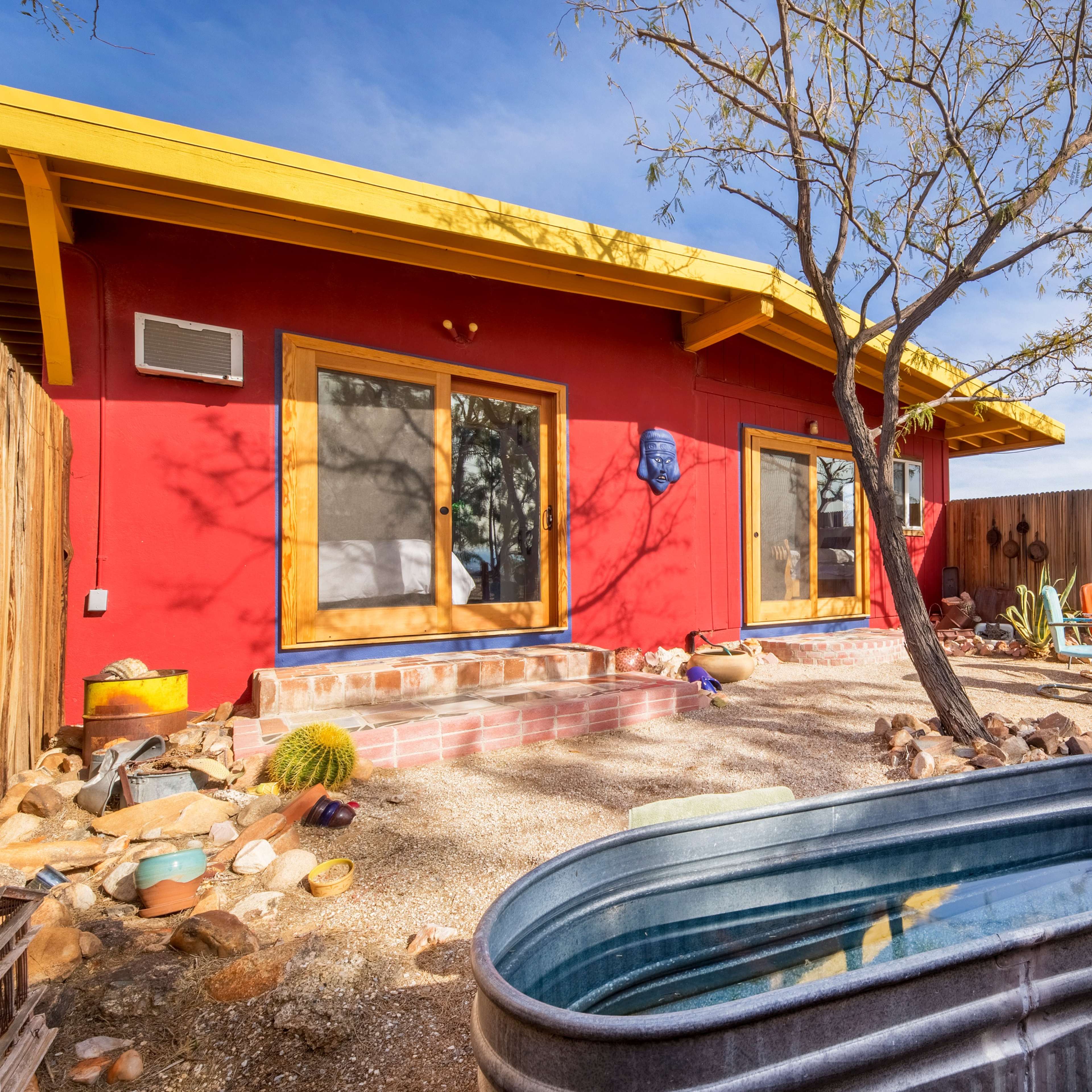 A red house with large windows and a yellow roof is situated in a desert landscape, featuring a metal tub and a few chairs in the foreground.