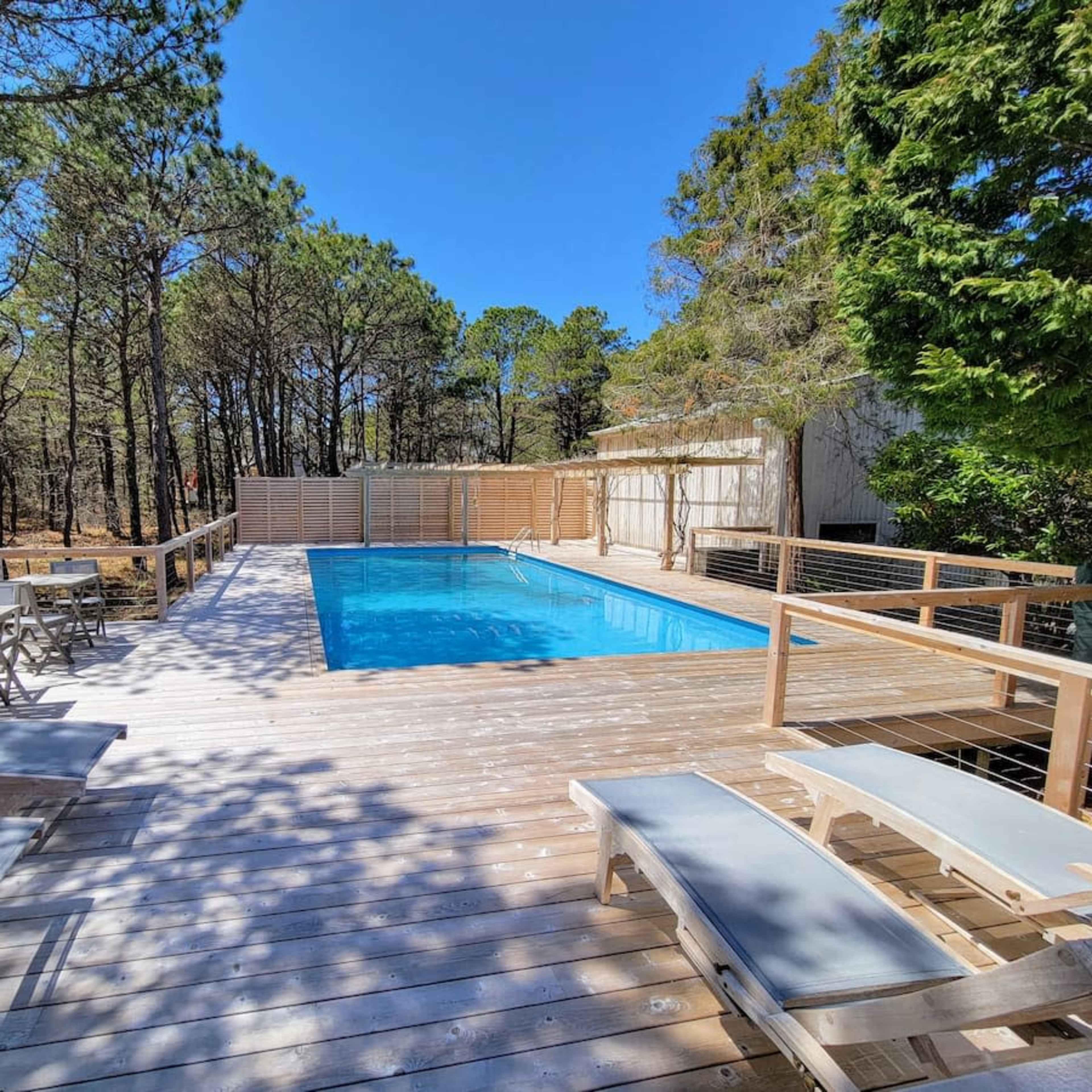 A rectangular pool surrounded by a wooden deck, with lounge chairs positioned nearby and trees in the background.