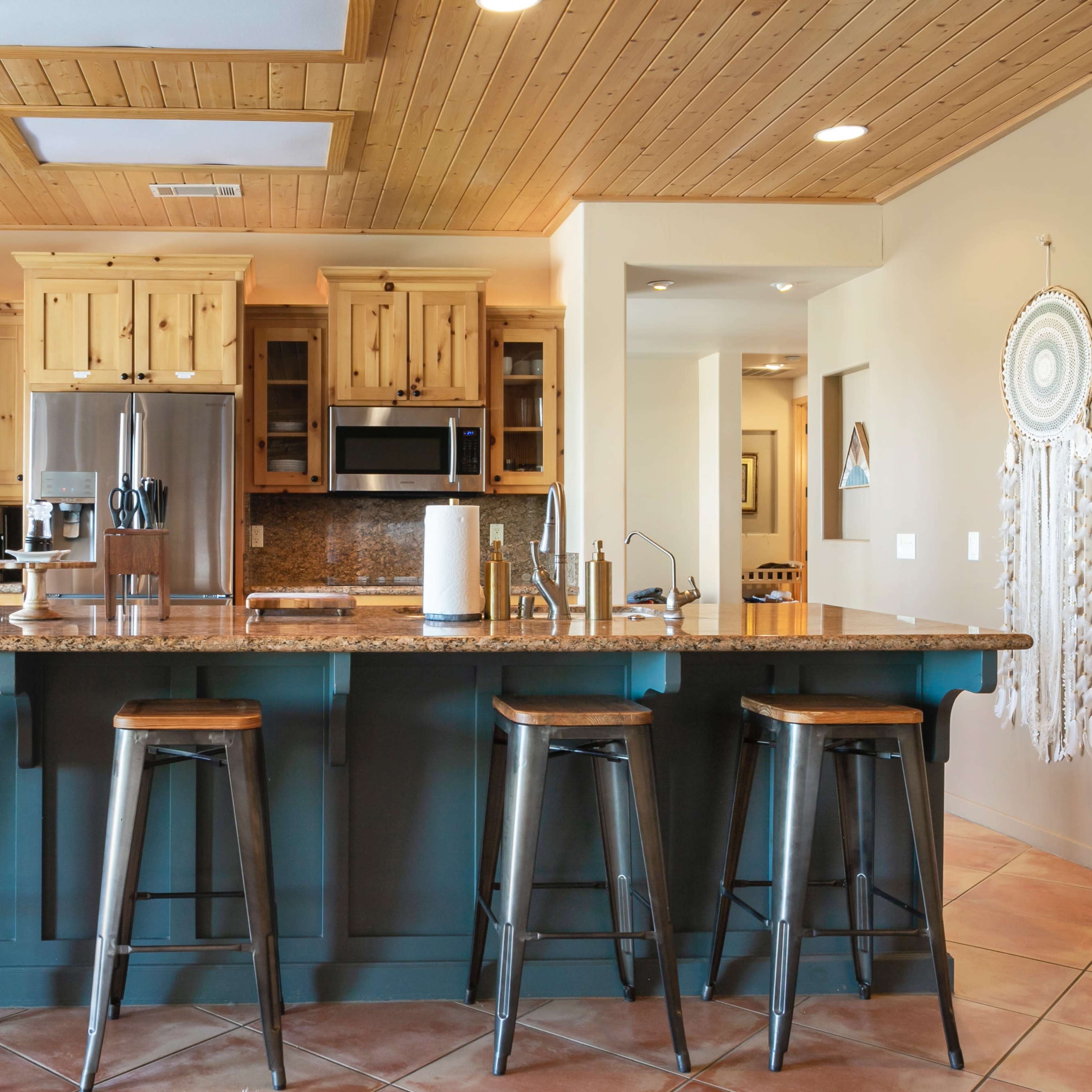 A modern kitchen with wooden cabinets, granite countertops, and four metal stools arranged at a central island.