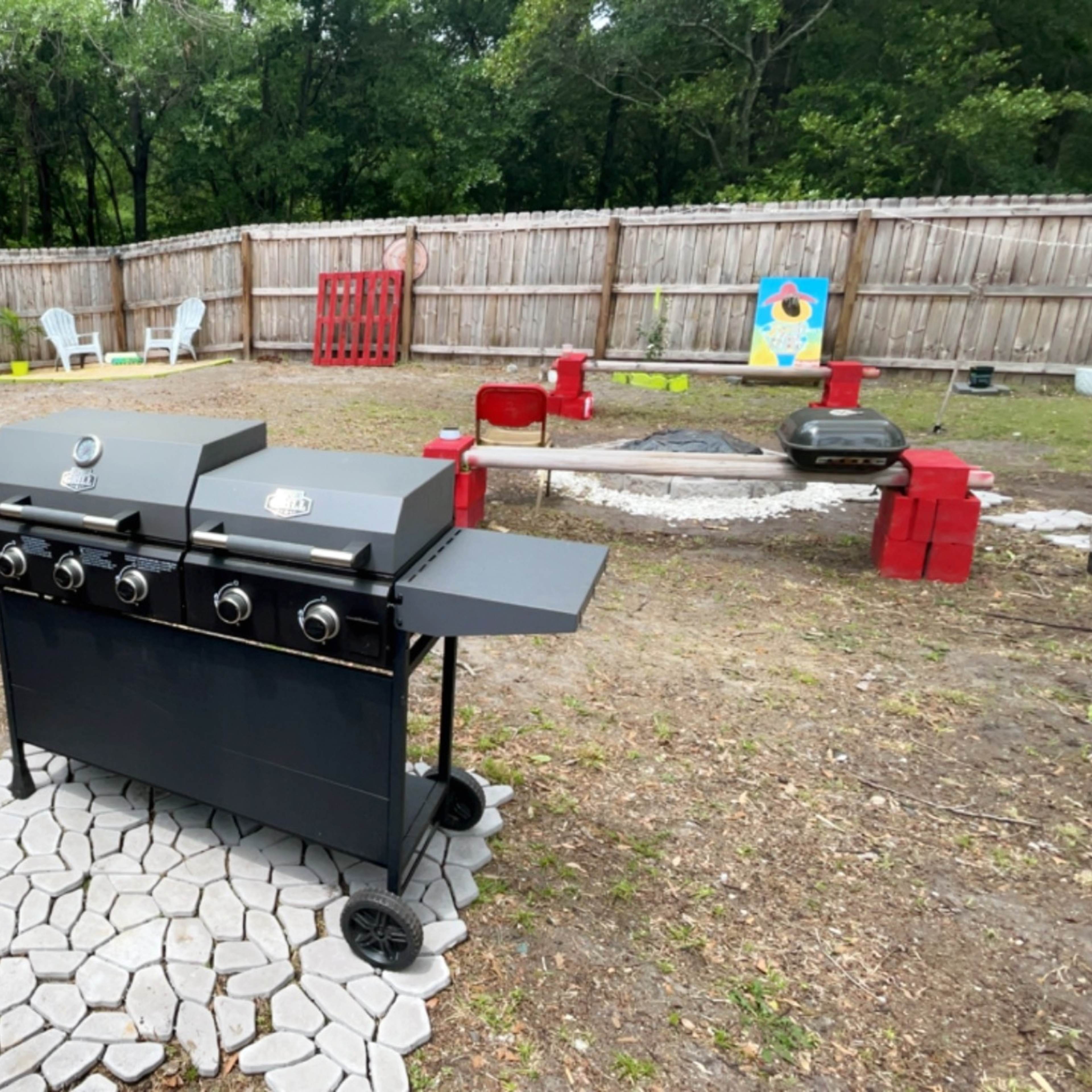 A black gas grill sits on a stone patio in an outdoor backyard, surrounded by a wooden fence and various lawn furniture.
