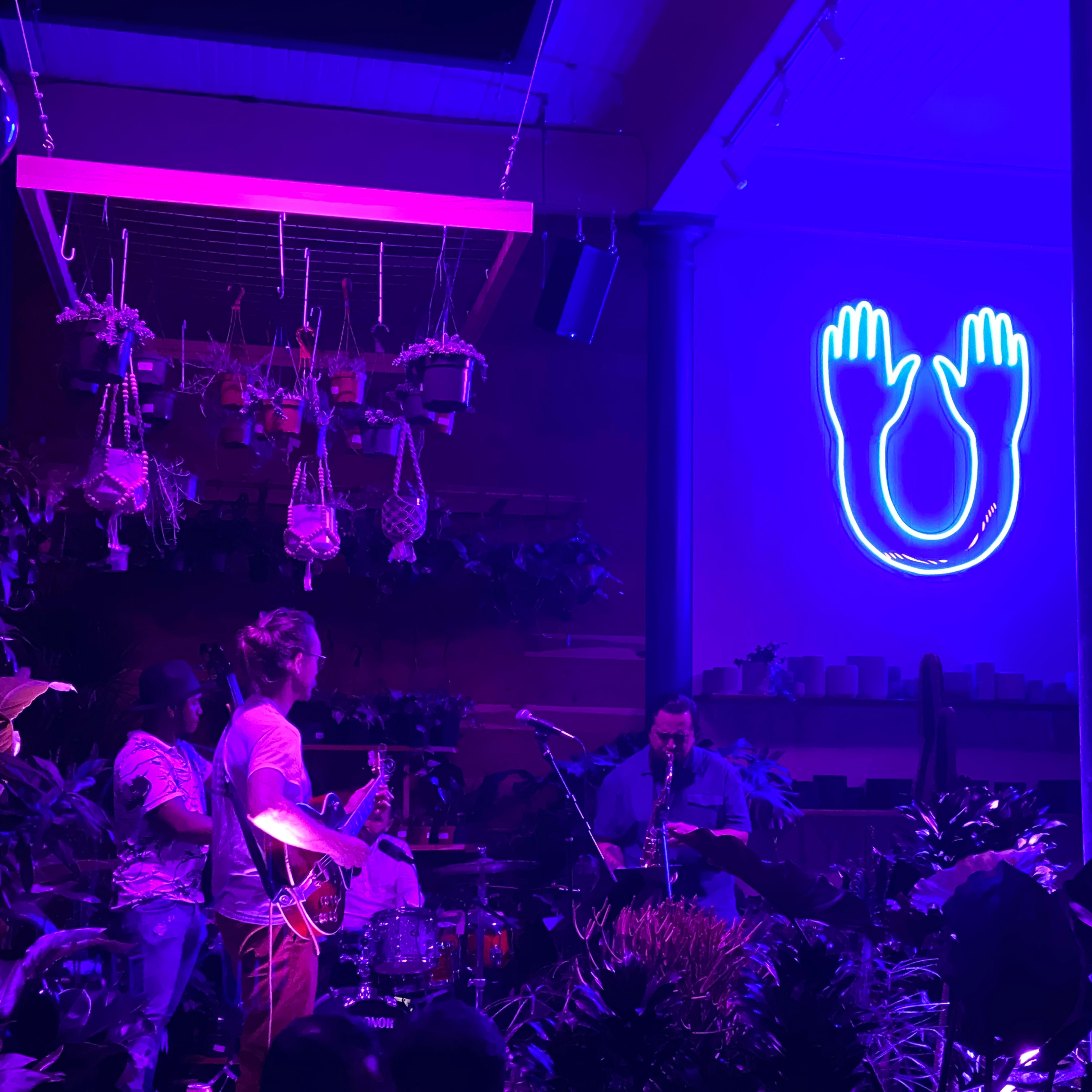 A band performs on stage in a dimly lit venue filled with plants, while a neon blue sign of hands and a horseshoe illuminates the background.