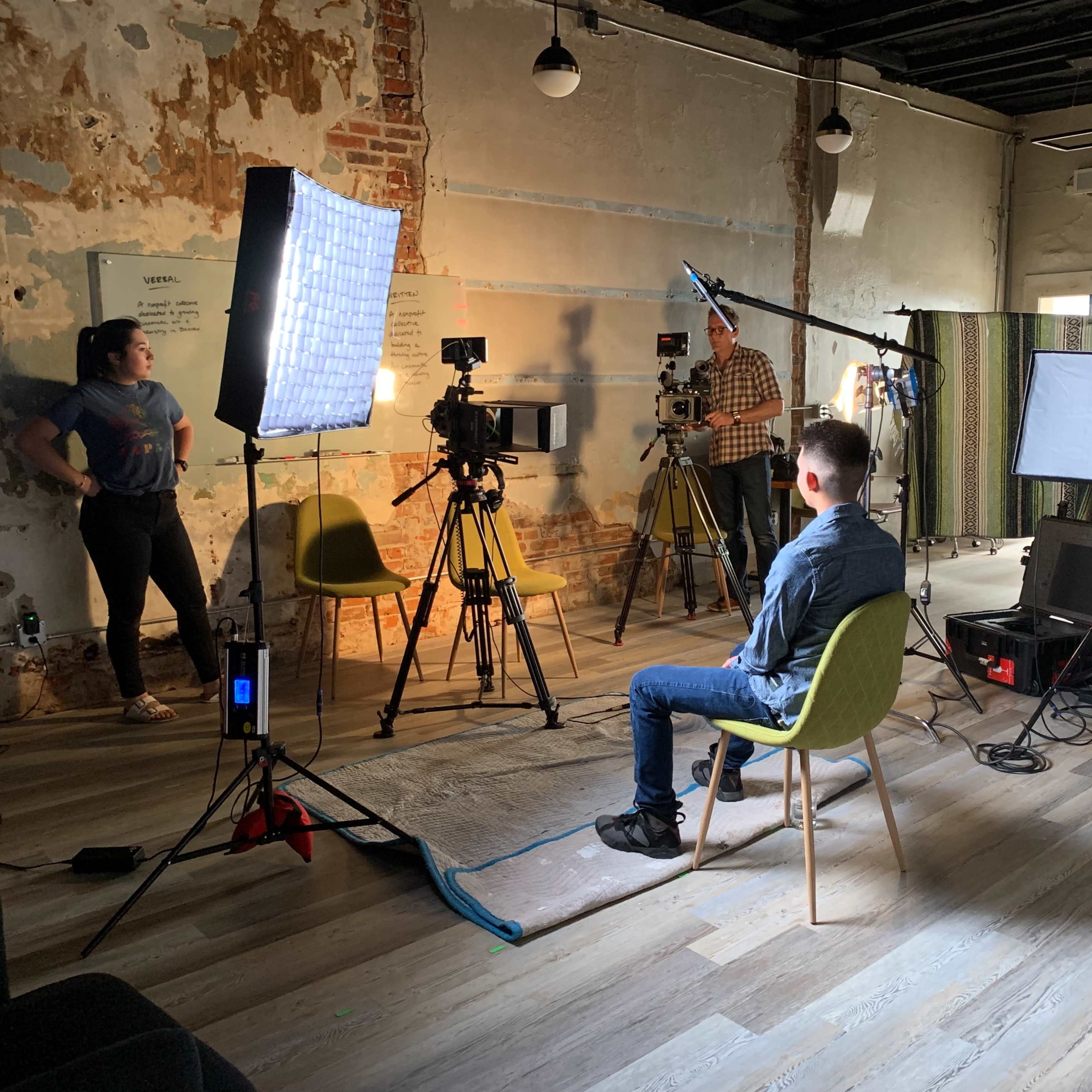 A film crew sets up equipment in a rustic room, preparing to shoot an interview with a person sitting on a chair.