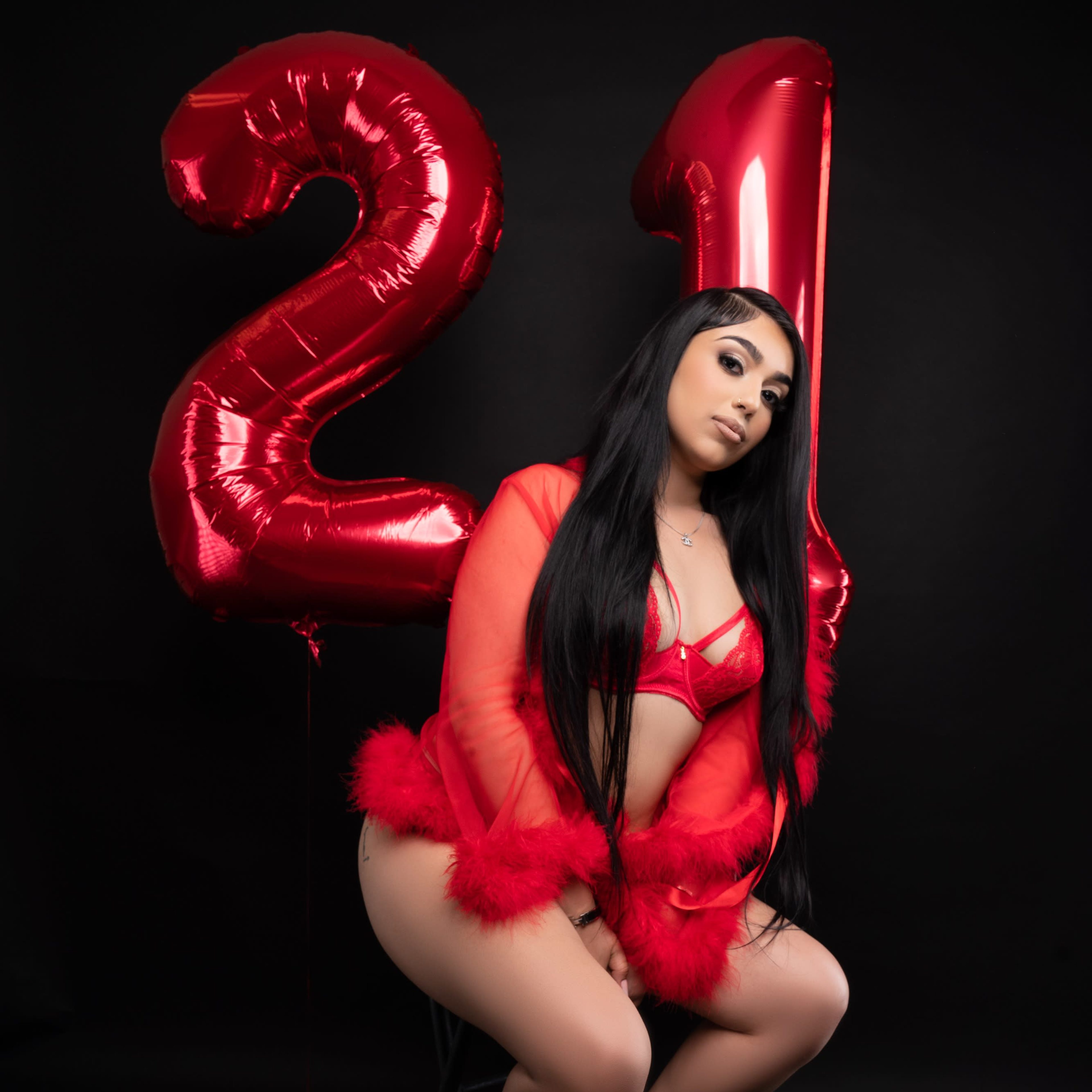 A woman in red lingerie sits on a stool next to large, red helium balloons that display the number "21" against a black background.