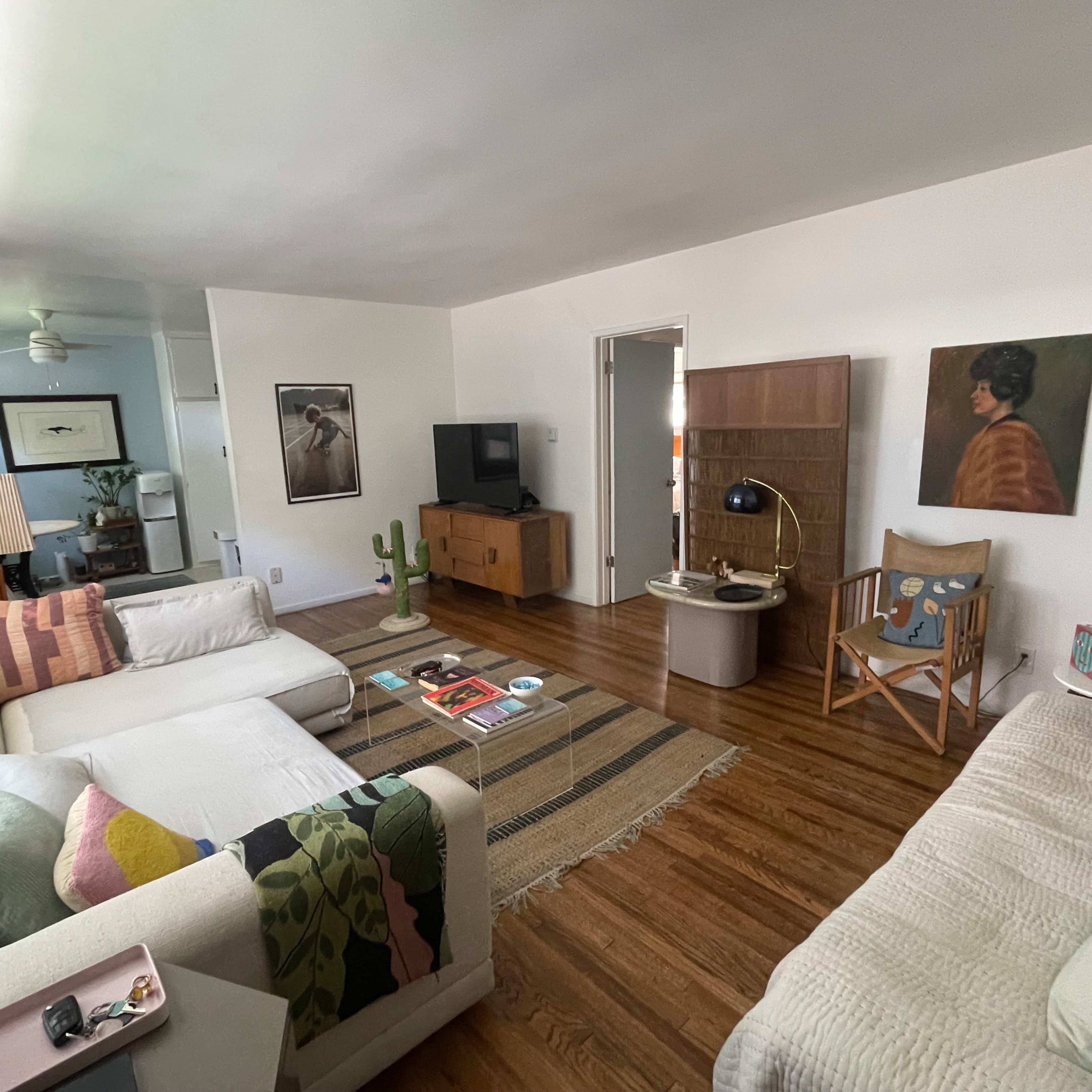 The living room features two sofas, a coffee table with magazines, a rug, and various decorative items, including a lamp and a cactus.