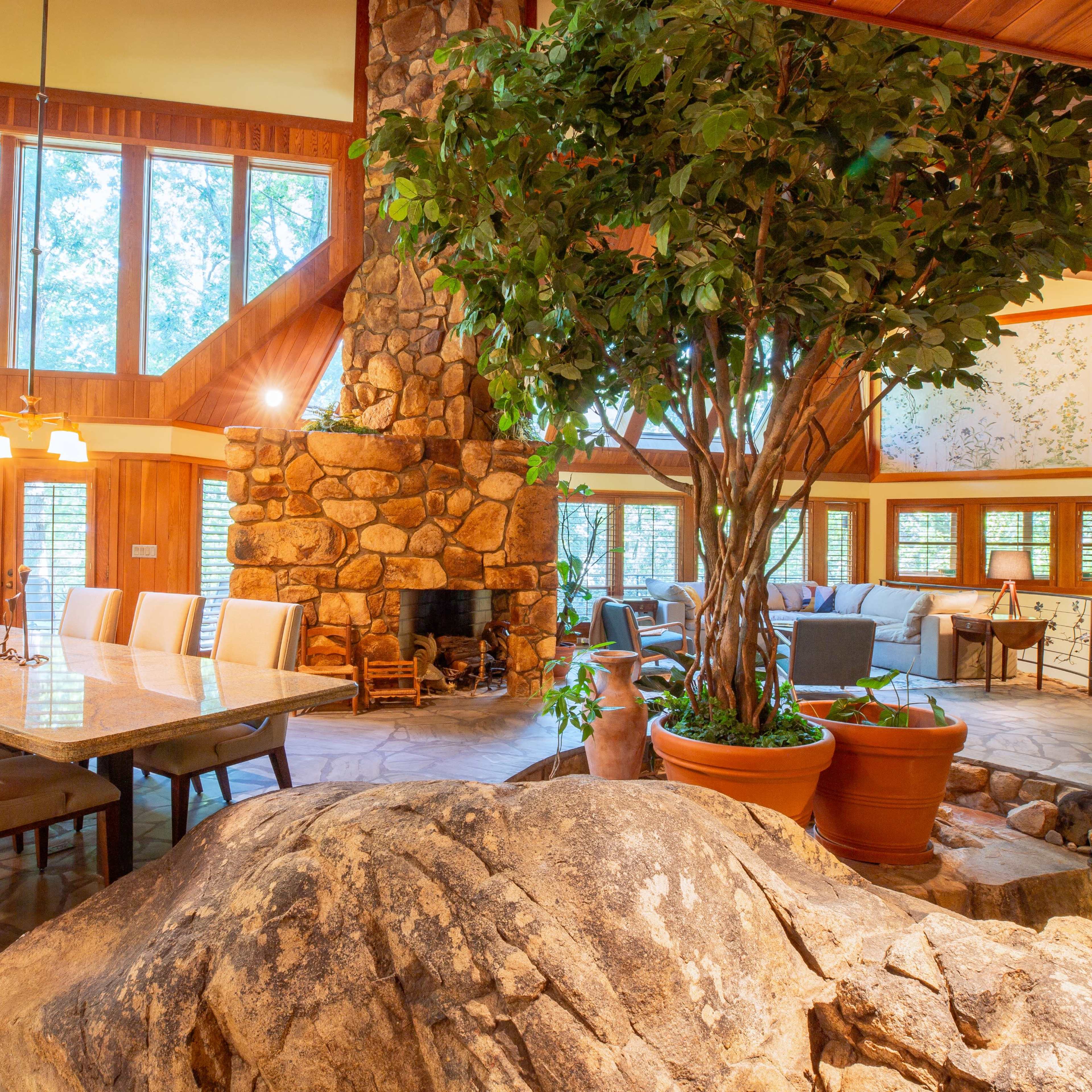 A spacious, rustic interior of a living area featuring a large stone fireplace, wooden beams, a dining table set for eight, and a decorative tree next to a large rock formation.