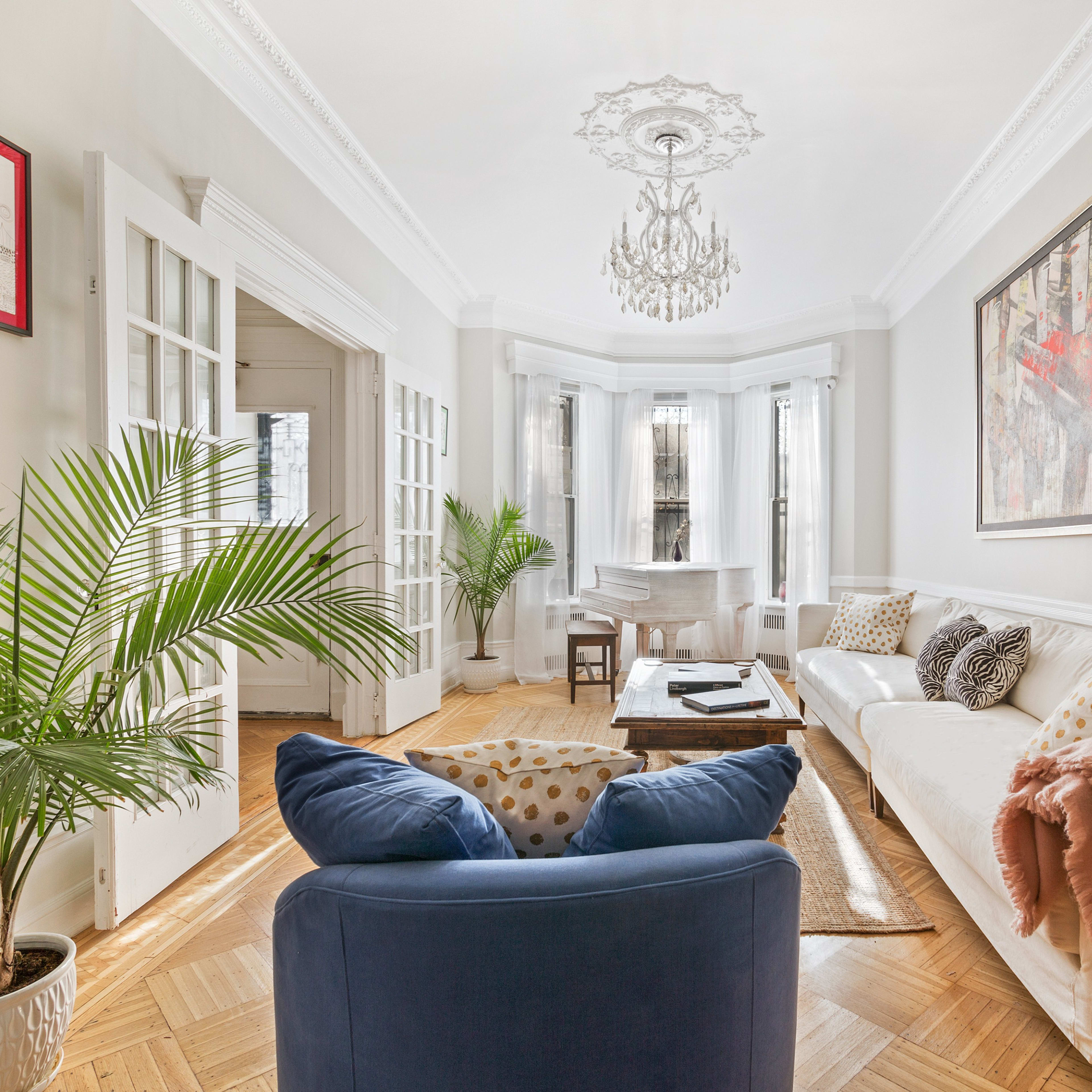 A spacious living room features a white sofa, a blue armchair, a piano in the corner, and large windows with natural light.