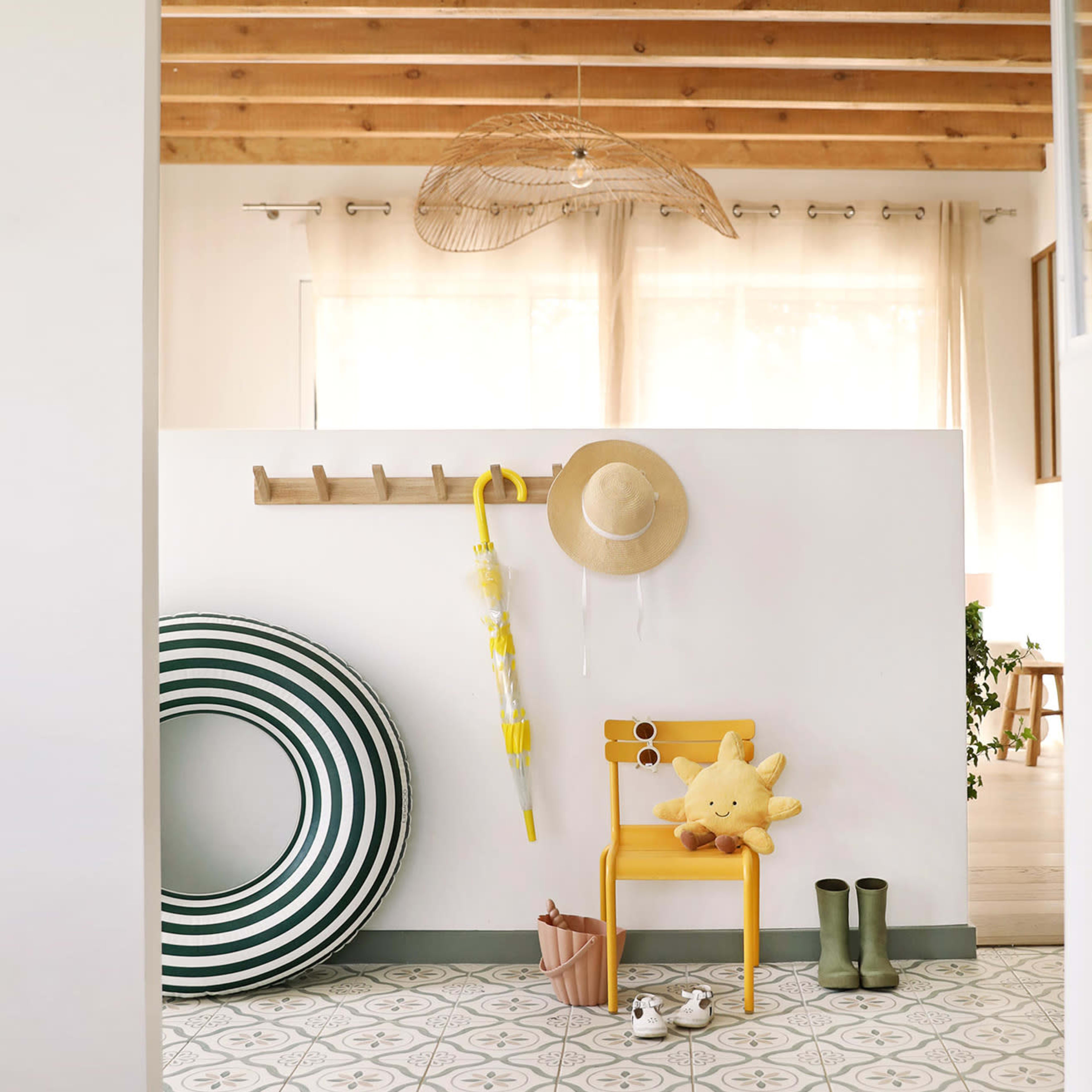 A bright entryway features a yellow chair adorned with a plush toy, a sun hat, and a yellow umbrella, beside a striped round mat and green rain boots.