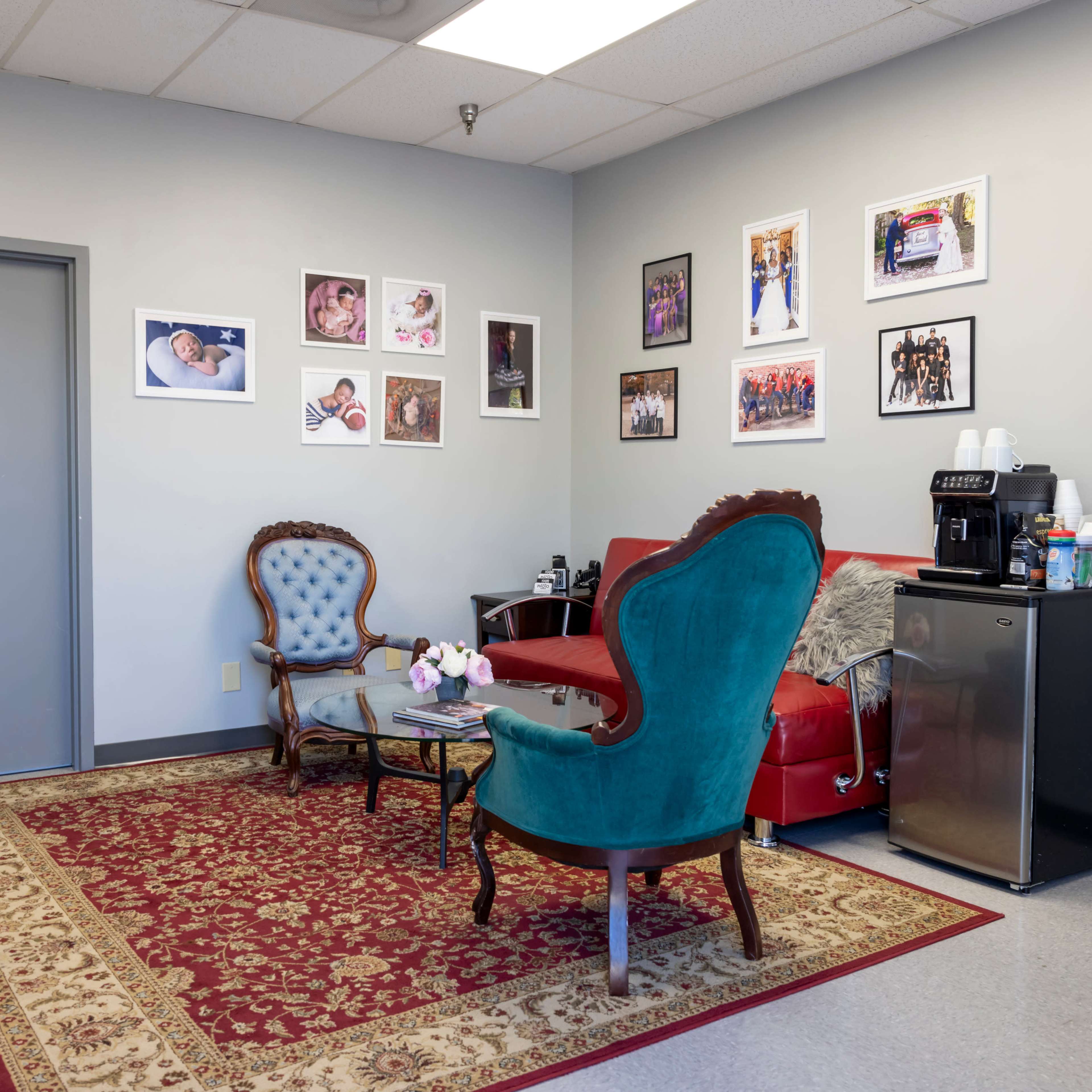 A waiting room with two upholstered chairs, a coffee station, and framed photographs on the wall above a red couch.