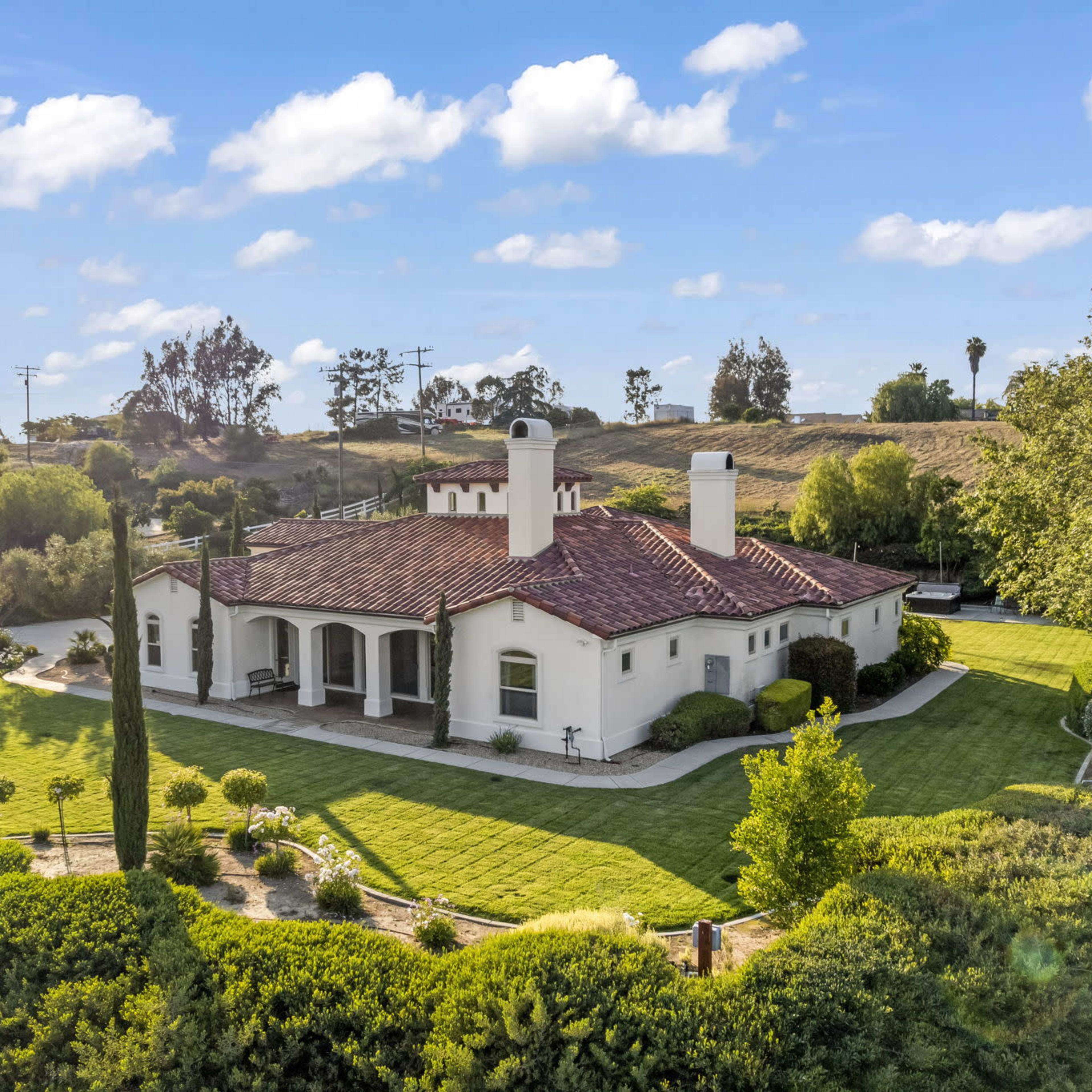 A single-story house with a tiled roof and manicured lawn is surrounded by trees and shrubs in a rural setting.