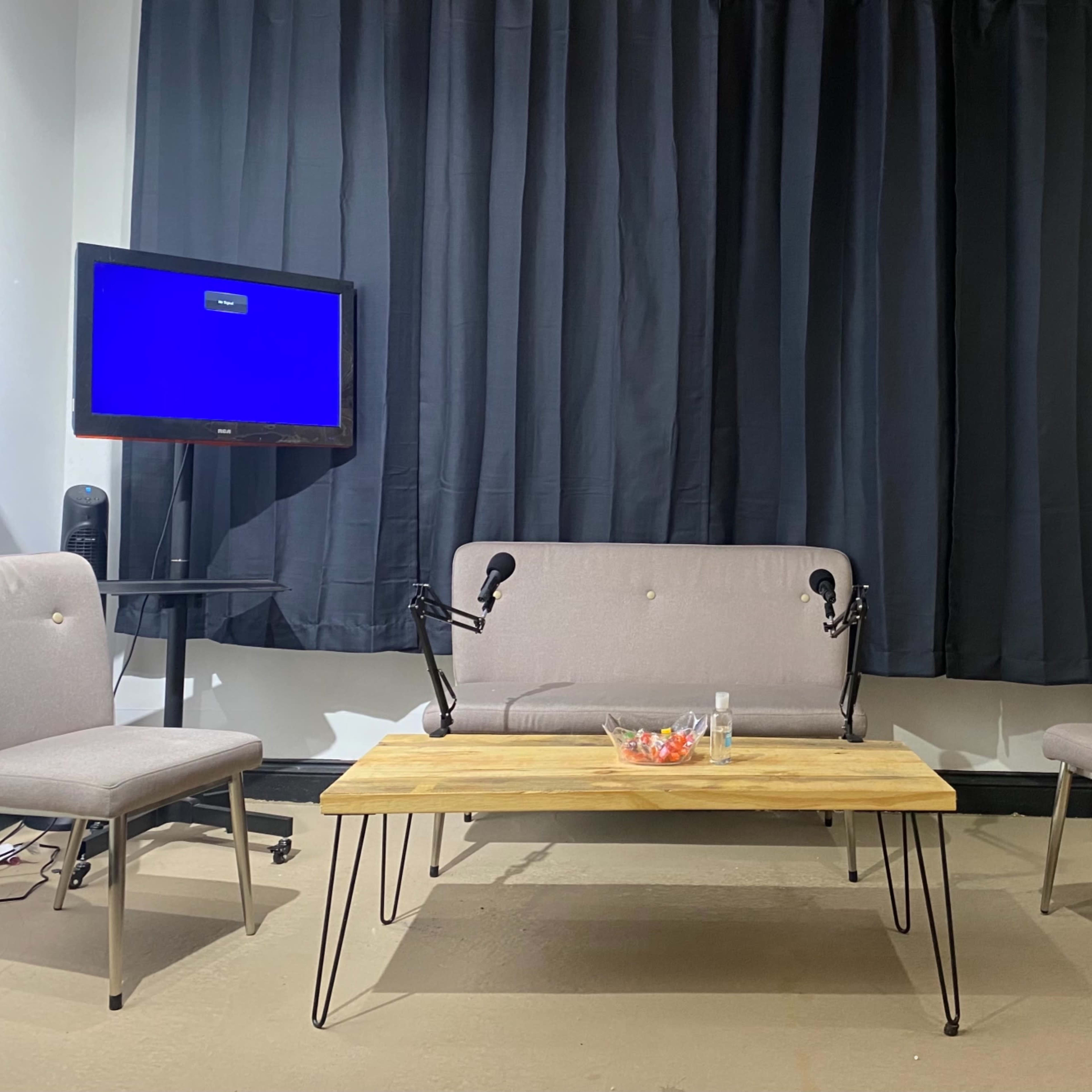 A small interview setup features a wooden table and two chairs, flanked by a couch, with a television displaying a blue screen in a brick-walled studio.