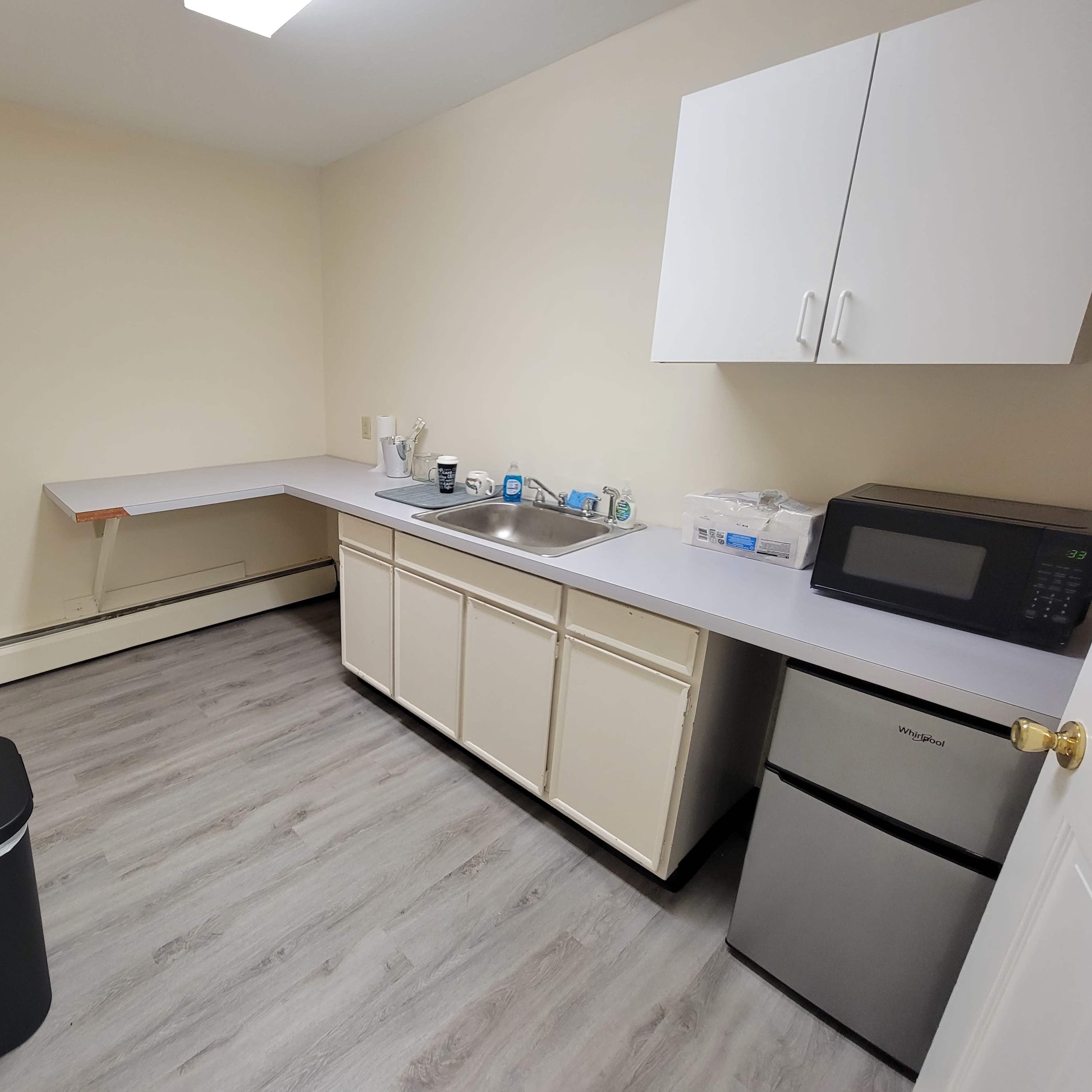 A small kitchen area with a sink, countertop, microwave, and a trash can, featuring white cabinets and light-colored flooring.