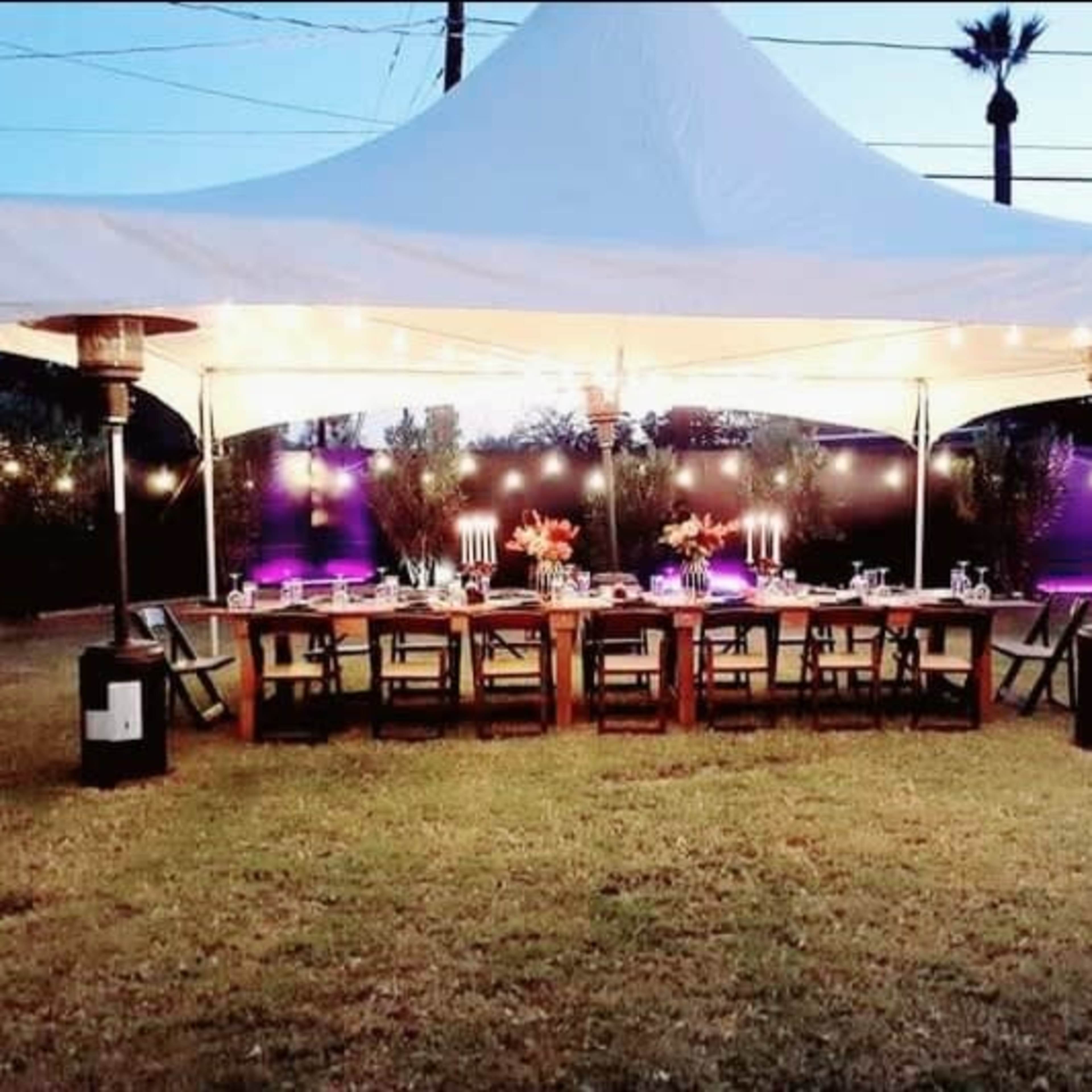 A large white tent is set up in a grassy area, featuring a long dining table surrounded by chairs, with decorative lights and plants in the background.