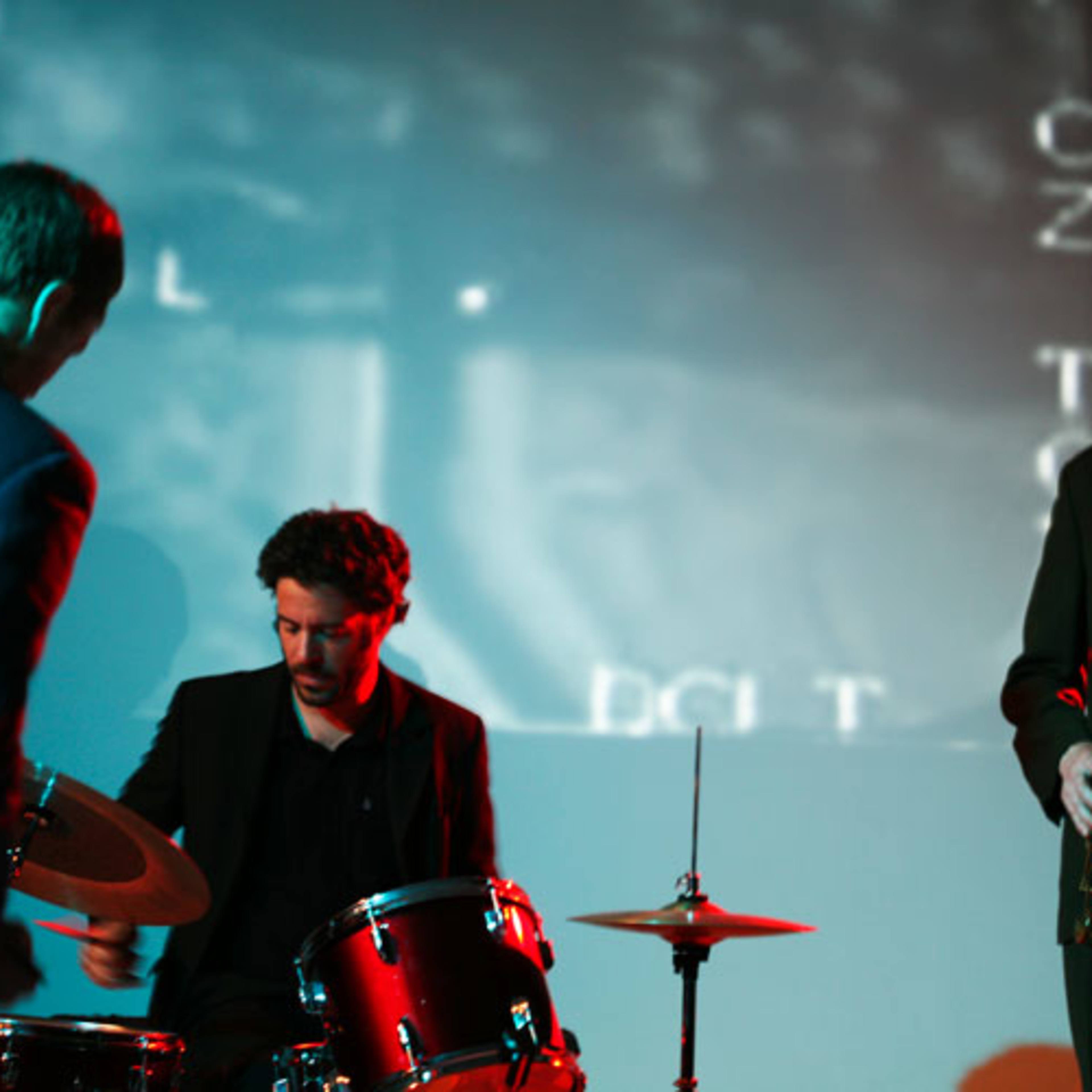 Three musicians perform on stage with a double bass, drums, and a saxophone, accompanied by a projected visual backdrop.