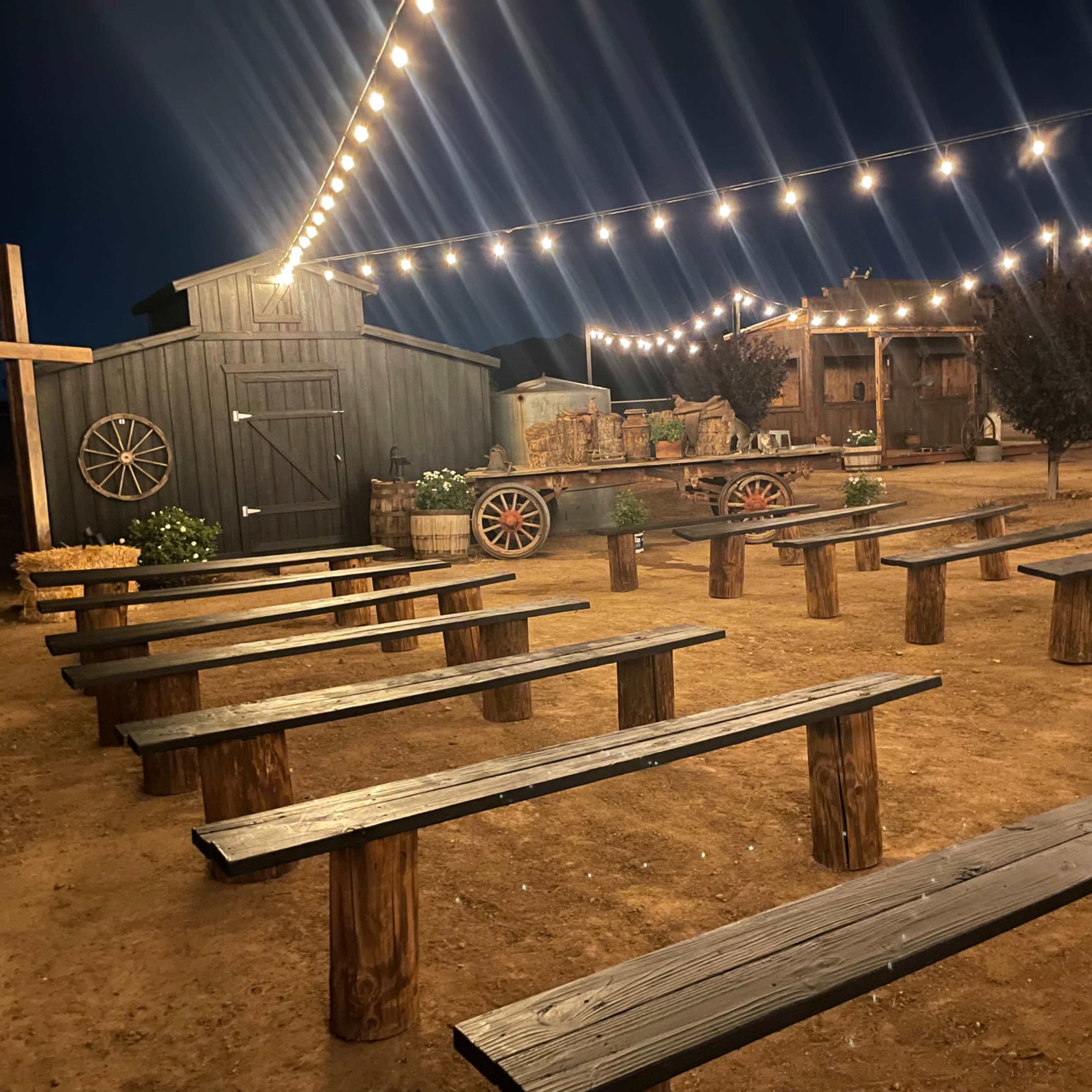 A rustic outdoor venue features wooden benches arranged in rows under string lights, with a barn and wagon in the background.