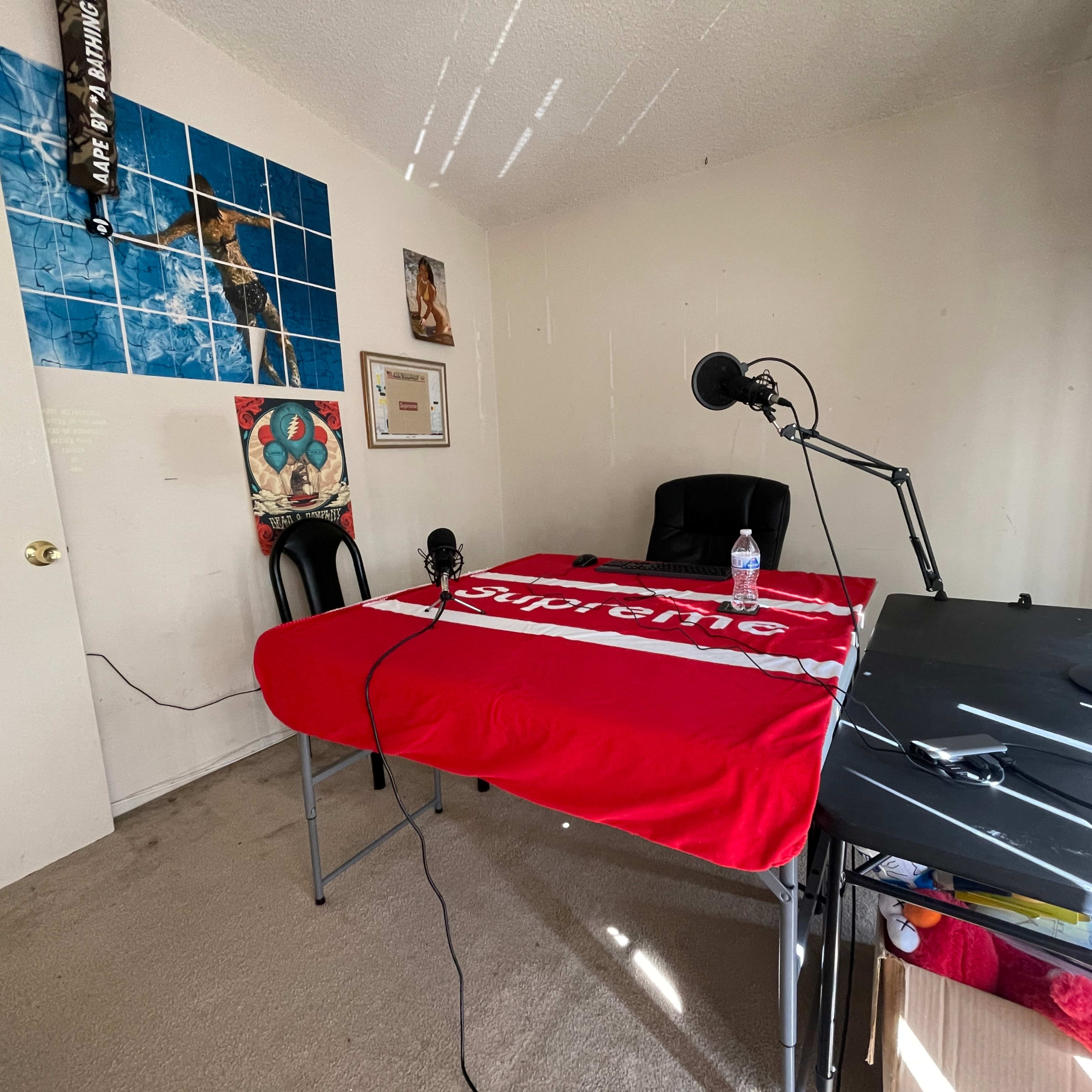 A small, sparsely furnished room with a table covered by a red Supreme blanket, two microphones set up on either side, a chair, and a desk with a monitor.