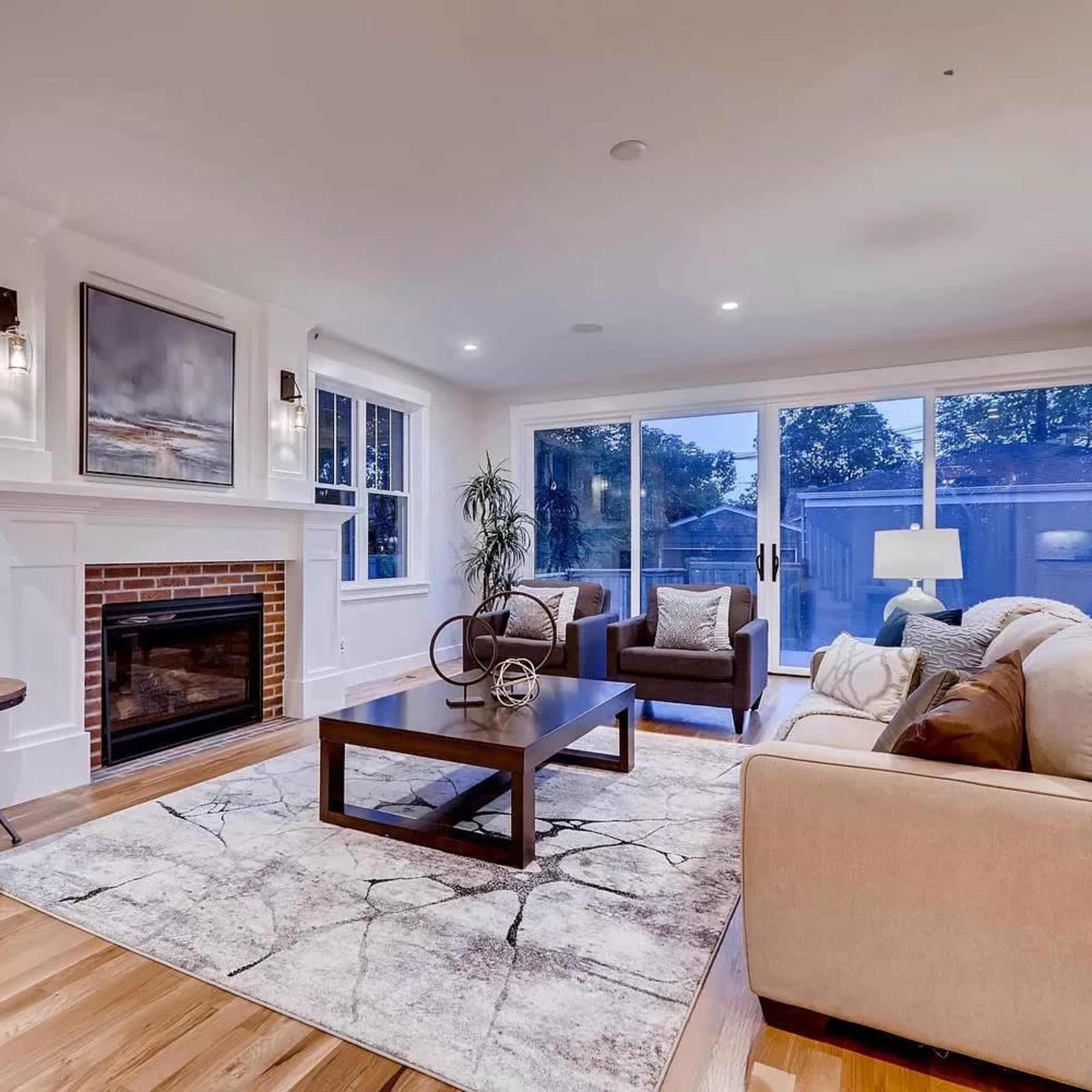 A modern living room with a fireplace, large windows, and a coffee table placed on a patterned area rug.