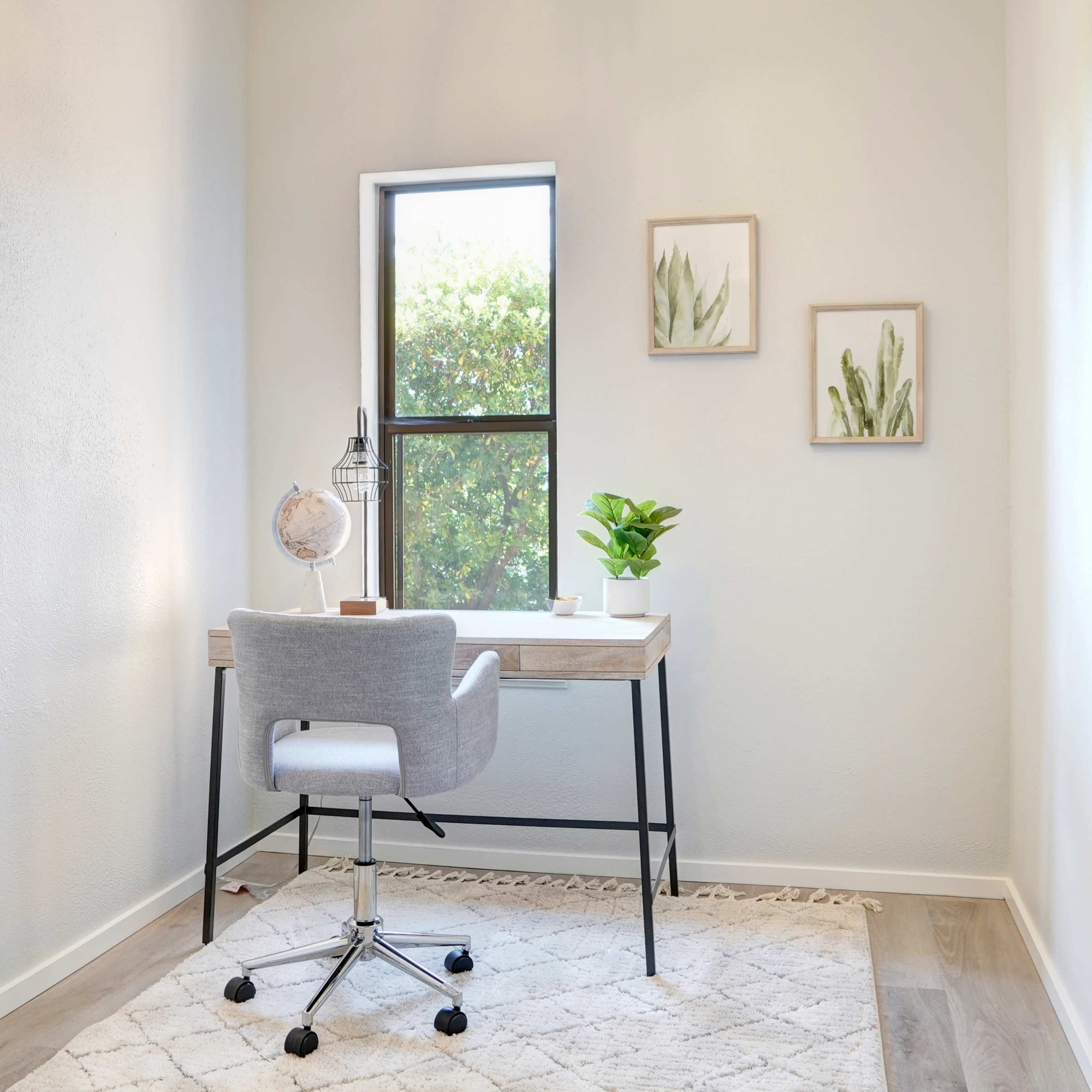 A small, well-lit home office featuring a wooden desk with a chair, a globe, a potted plant, and framed artwork on the walls.