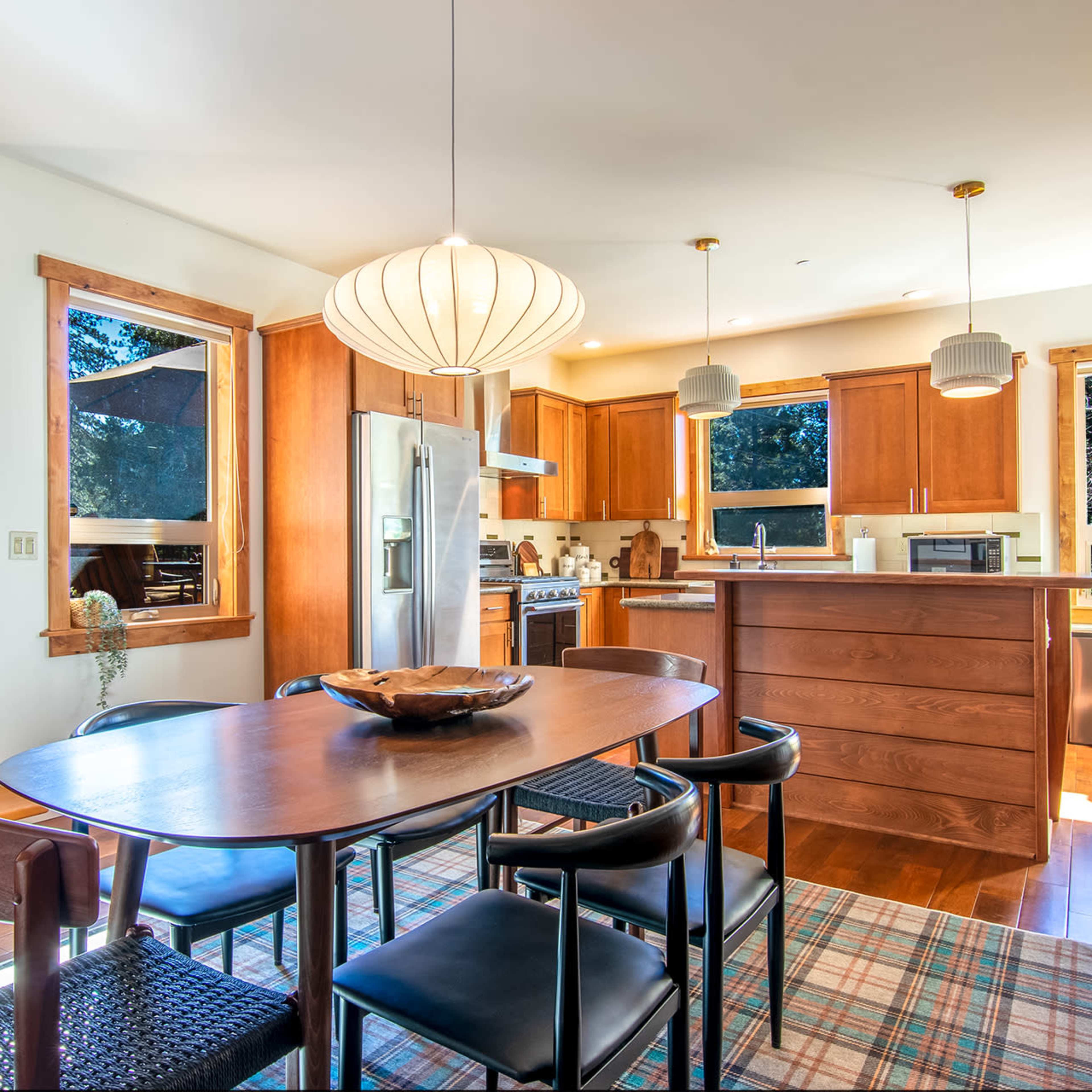 A dining area features a round table surrounded by black chairs, positioned near a well-lit kitchen with wooden cabinets and large windows.