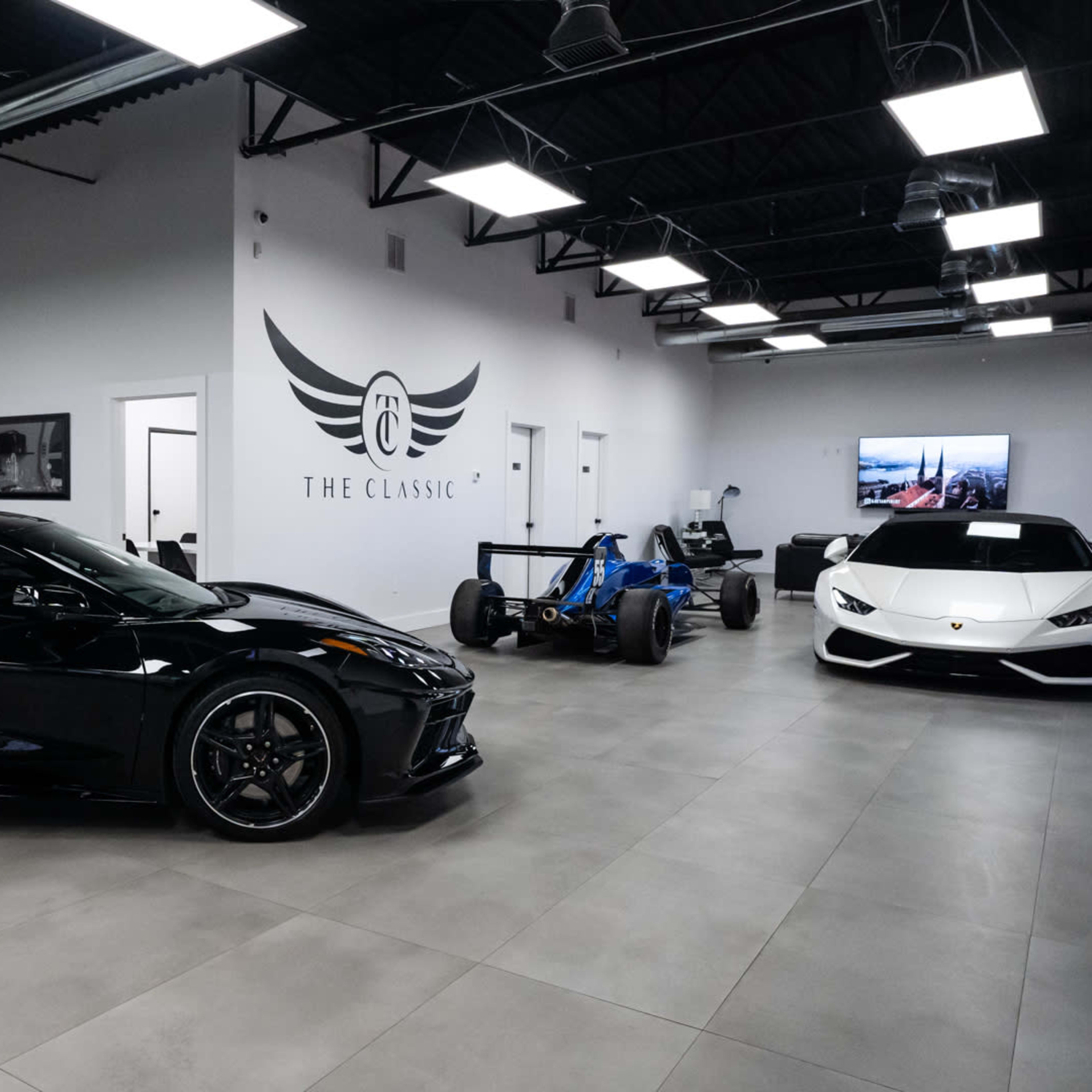 A sleek car showroom featuring a black sports car, a blue racing car, and a white luxury vehicle, all displayed under bright lights in a modern, clean space.