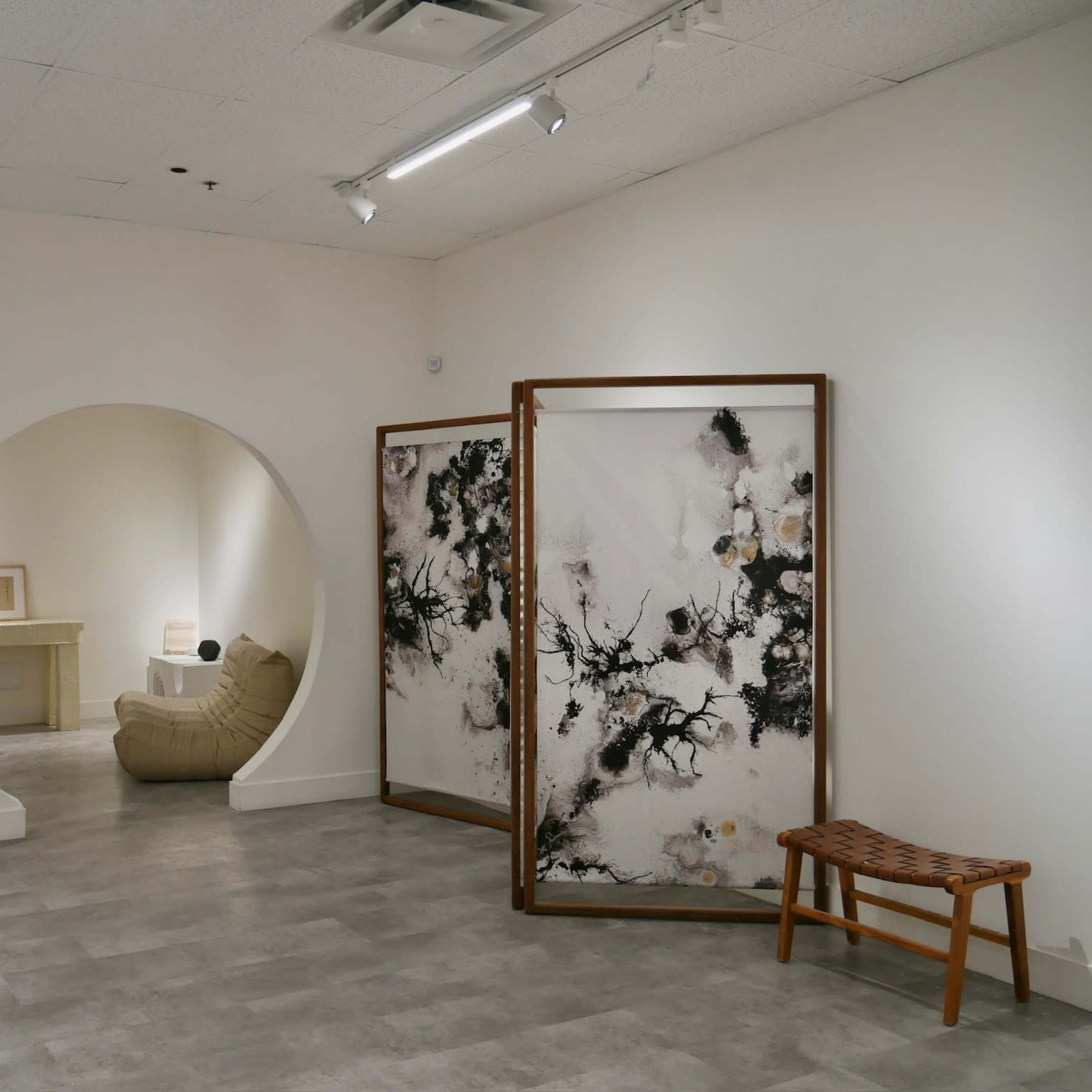 A minimalist interior space with a curved archway, a beige chair, a decorative folding screen featuring abstract art, and wooden furniture.