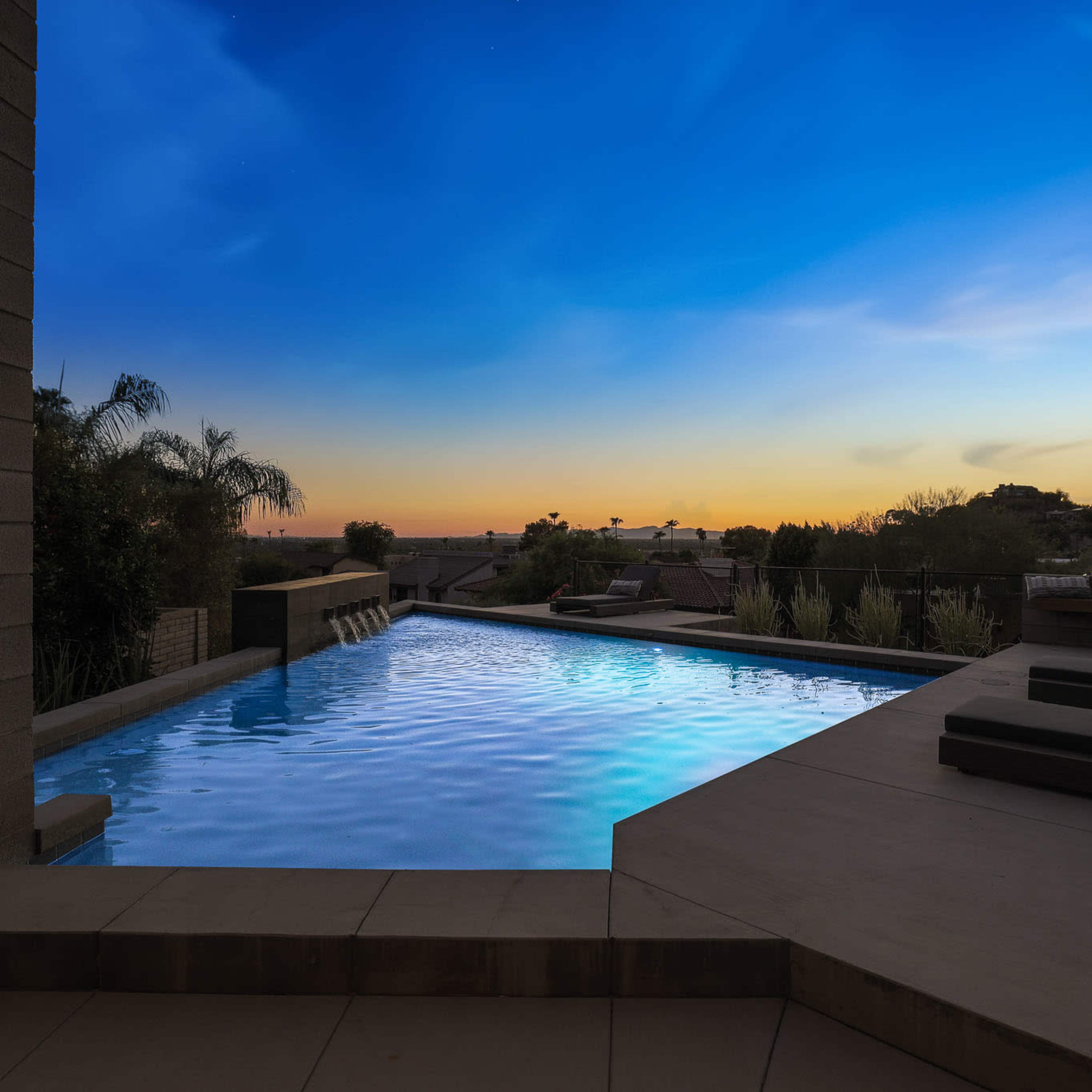 A contemporary swimming pool with an infinity edge, set against a twilight sky and overlooking a quiet suburban landscape.
