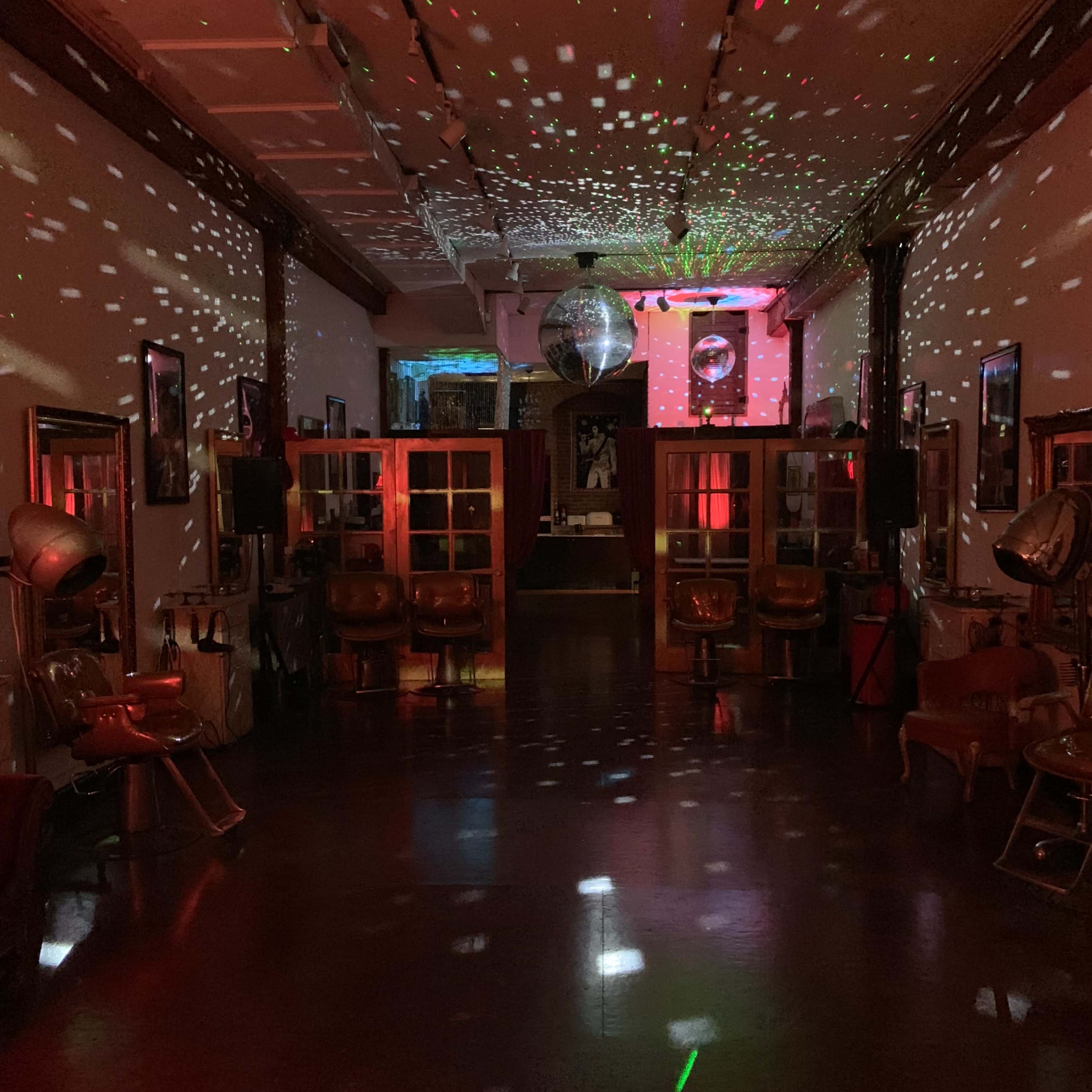 A dimly lit salon with red and green lights creating a disco effect, featuring vintage salon chairs and a large disco ball suspended from the ceiling.