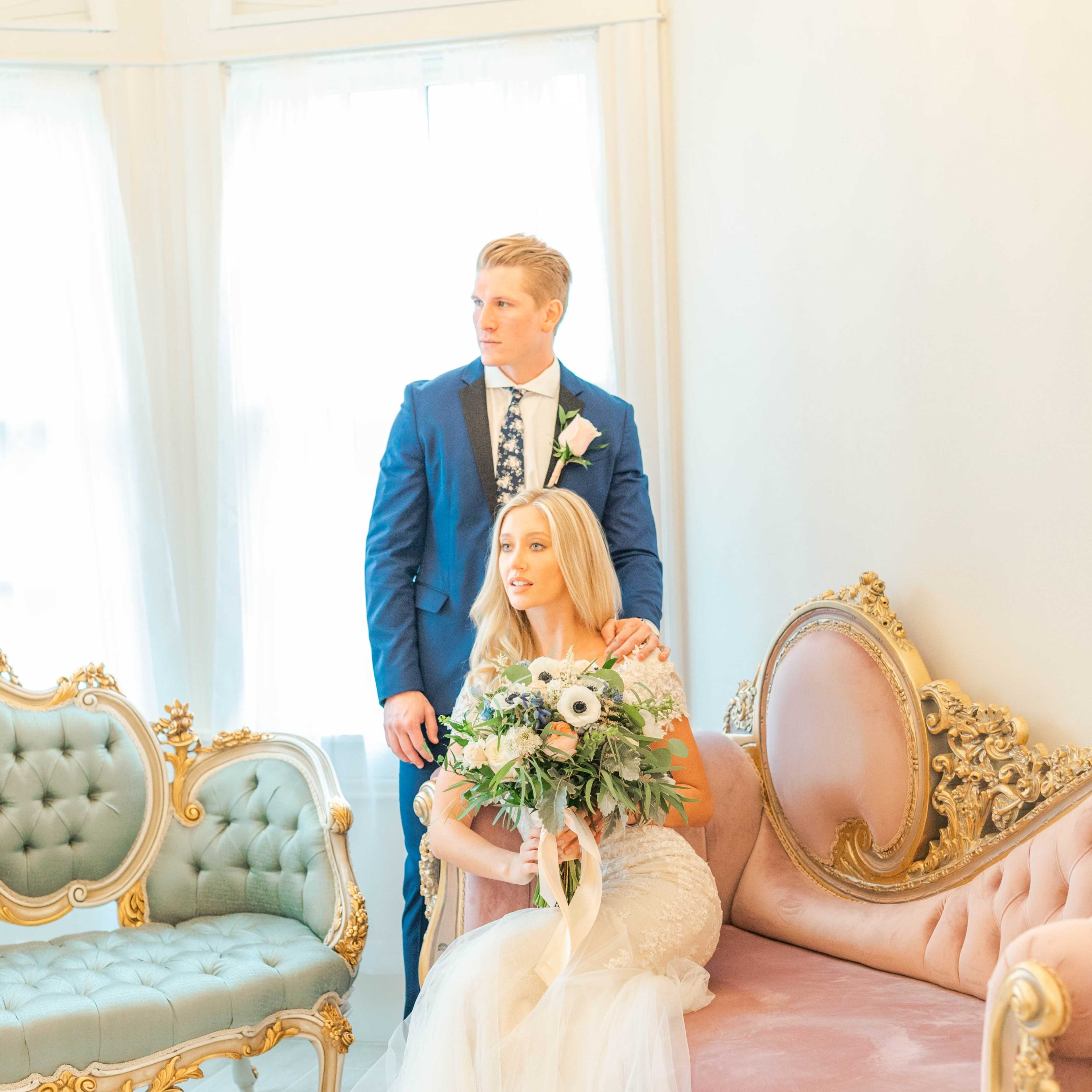 A man in a blue suit stands behind a woman holding a bouquet, seated on a pink couch in a brightly lit room with elegant decor.