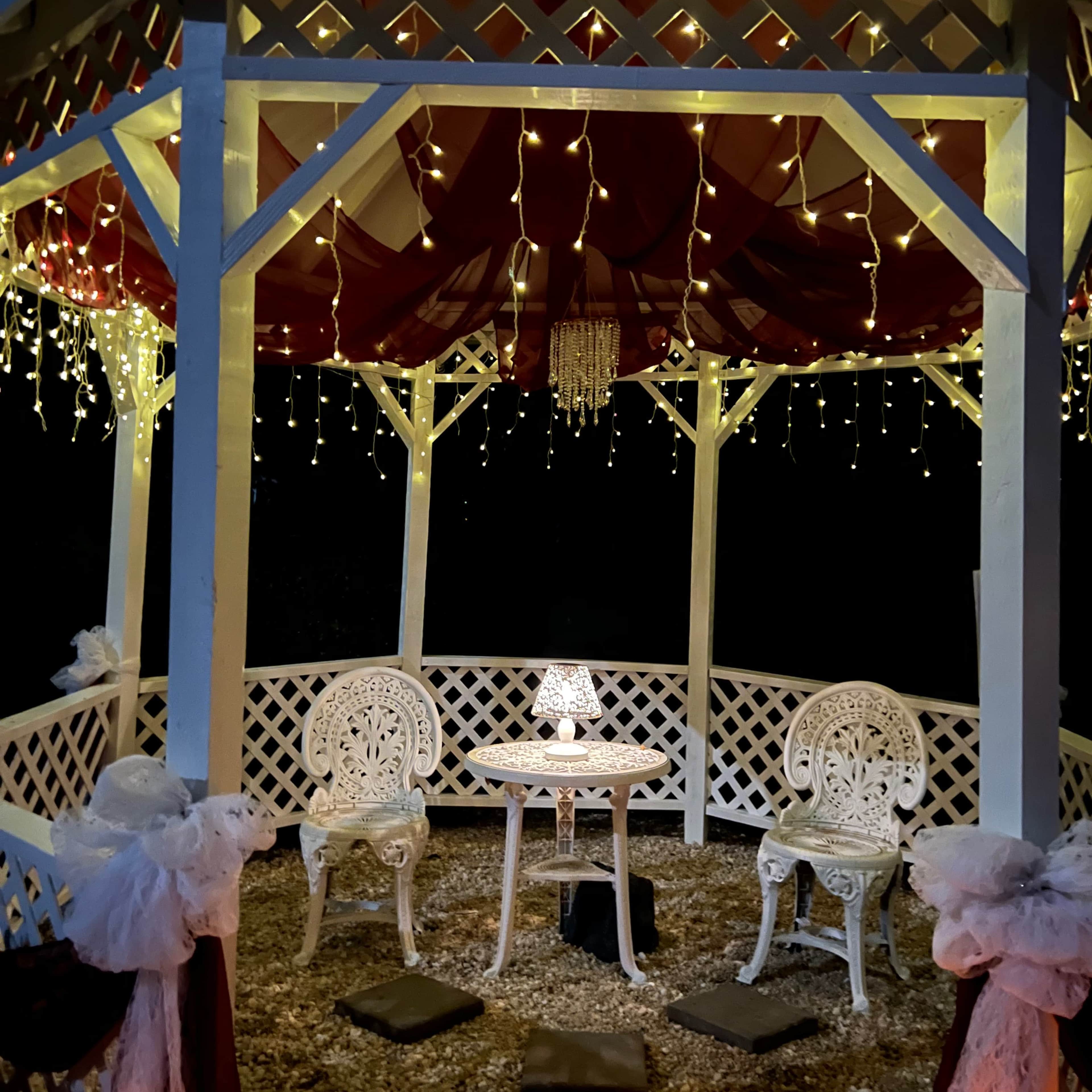 A gazebo is illuminated with string lights and features a small table and two chairs on a gravel floor.
