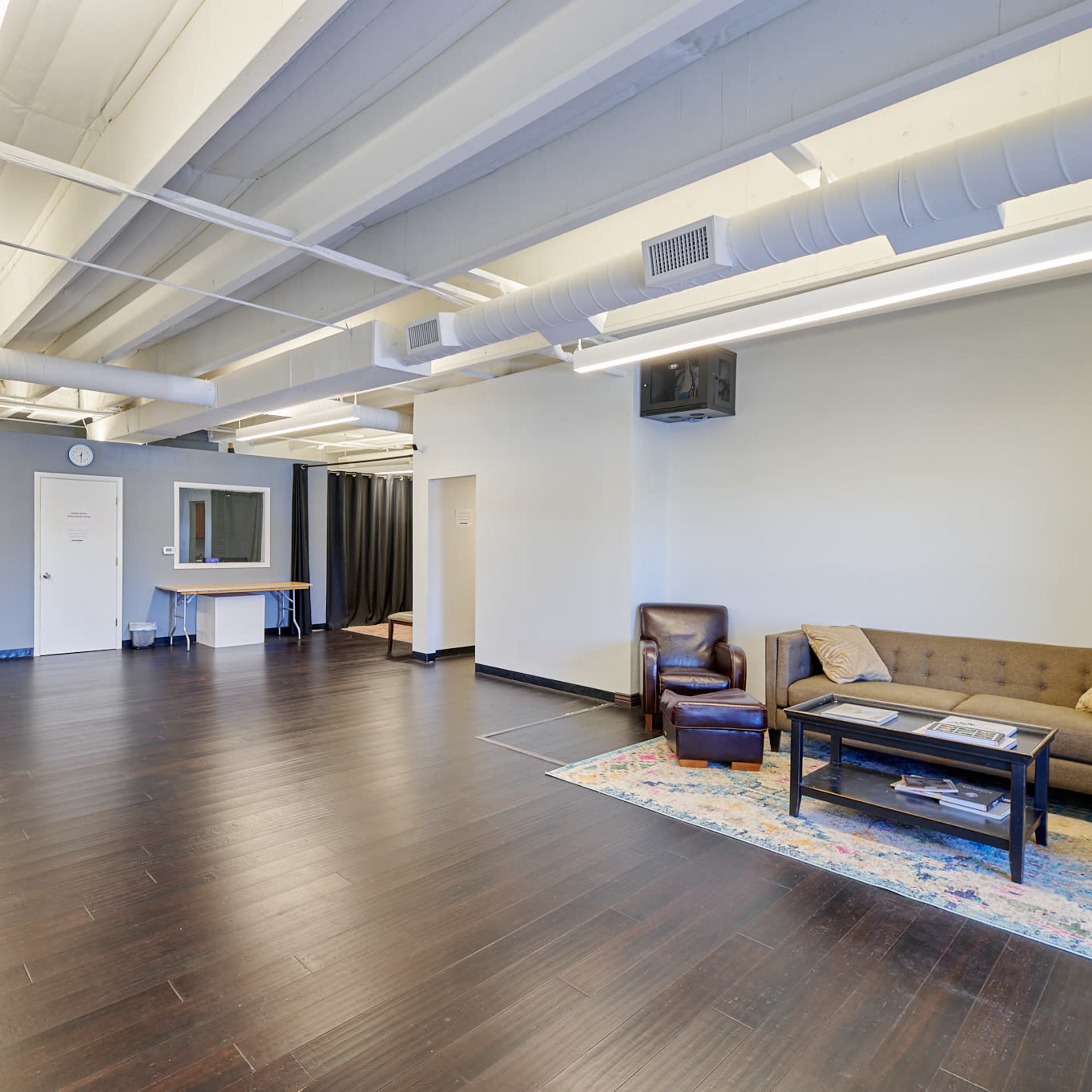 A spacious, unfurnished room with dark wooden floors, a couch, a coffee table, and a small kitchenette area.