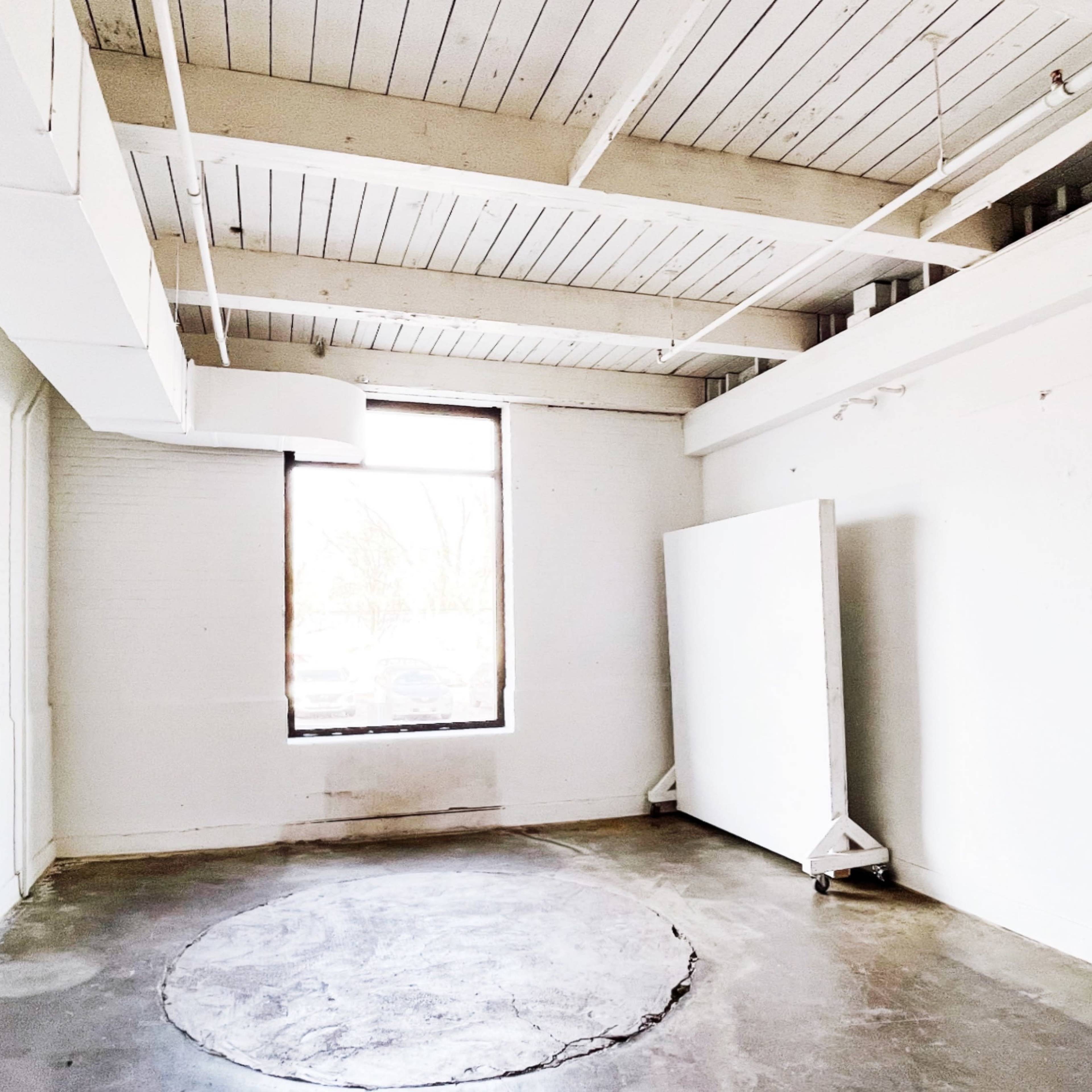 A spacious, empty room with white walls, a circular concrete area on the floor, and a large window letting in natural light.