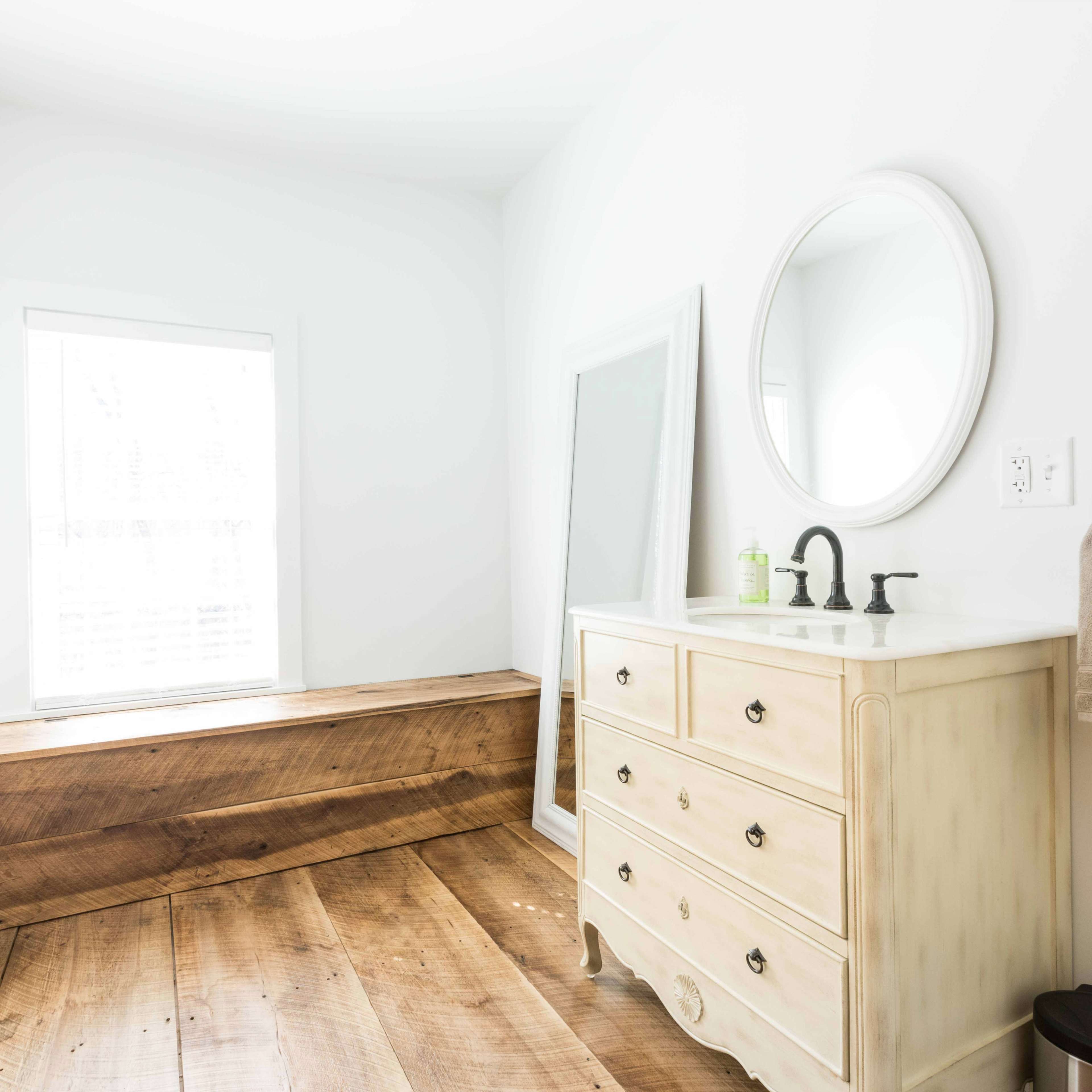 A bright bathroom featuring a wooden floor, a beige vanity with a sink, a round mirror, and a window beside a wooden bench.