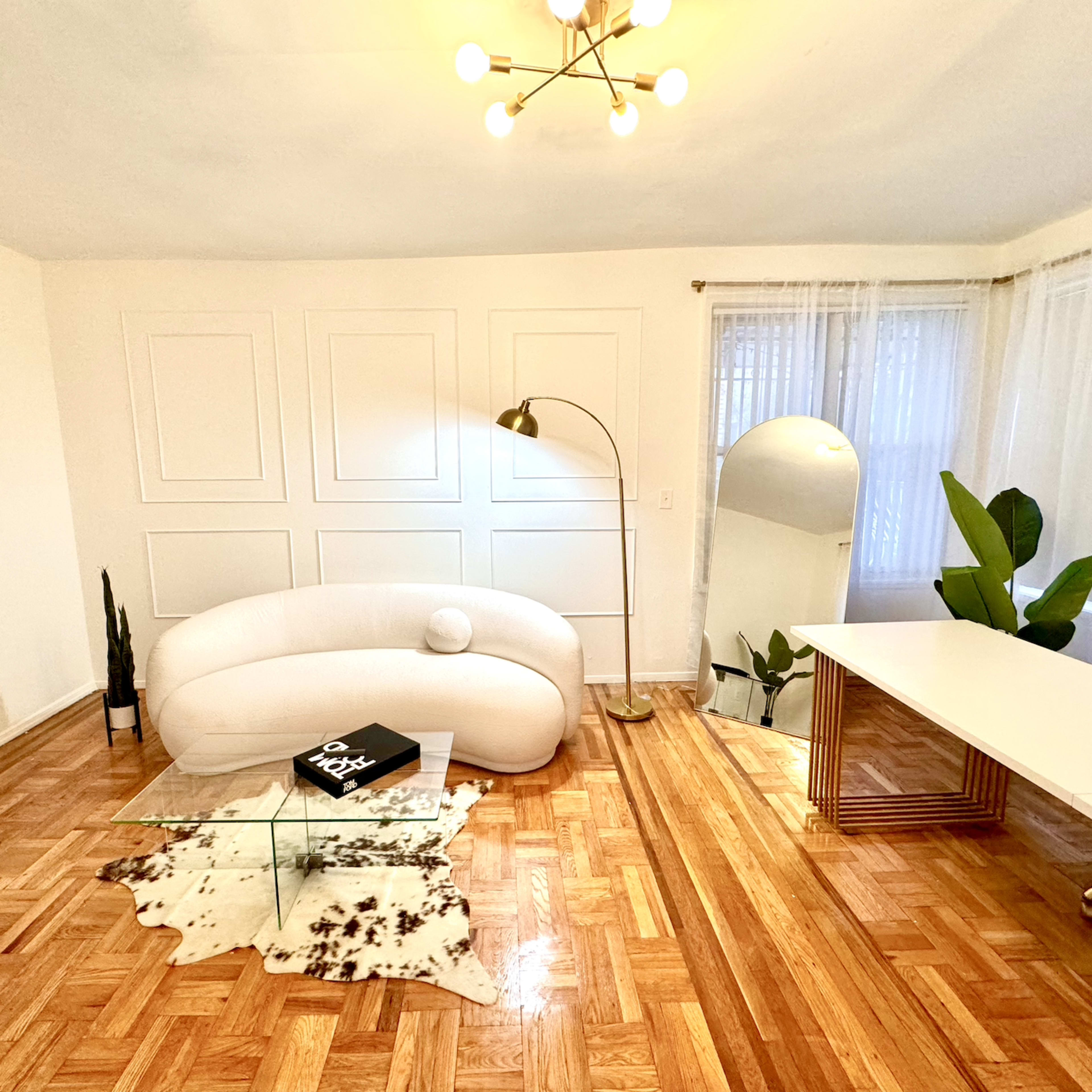 A modern, minimalist living space features a curved white sofa, a glass coffee table on a cowhide rug, a workspace with a desk, a large mirror, and potted plants.