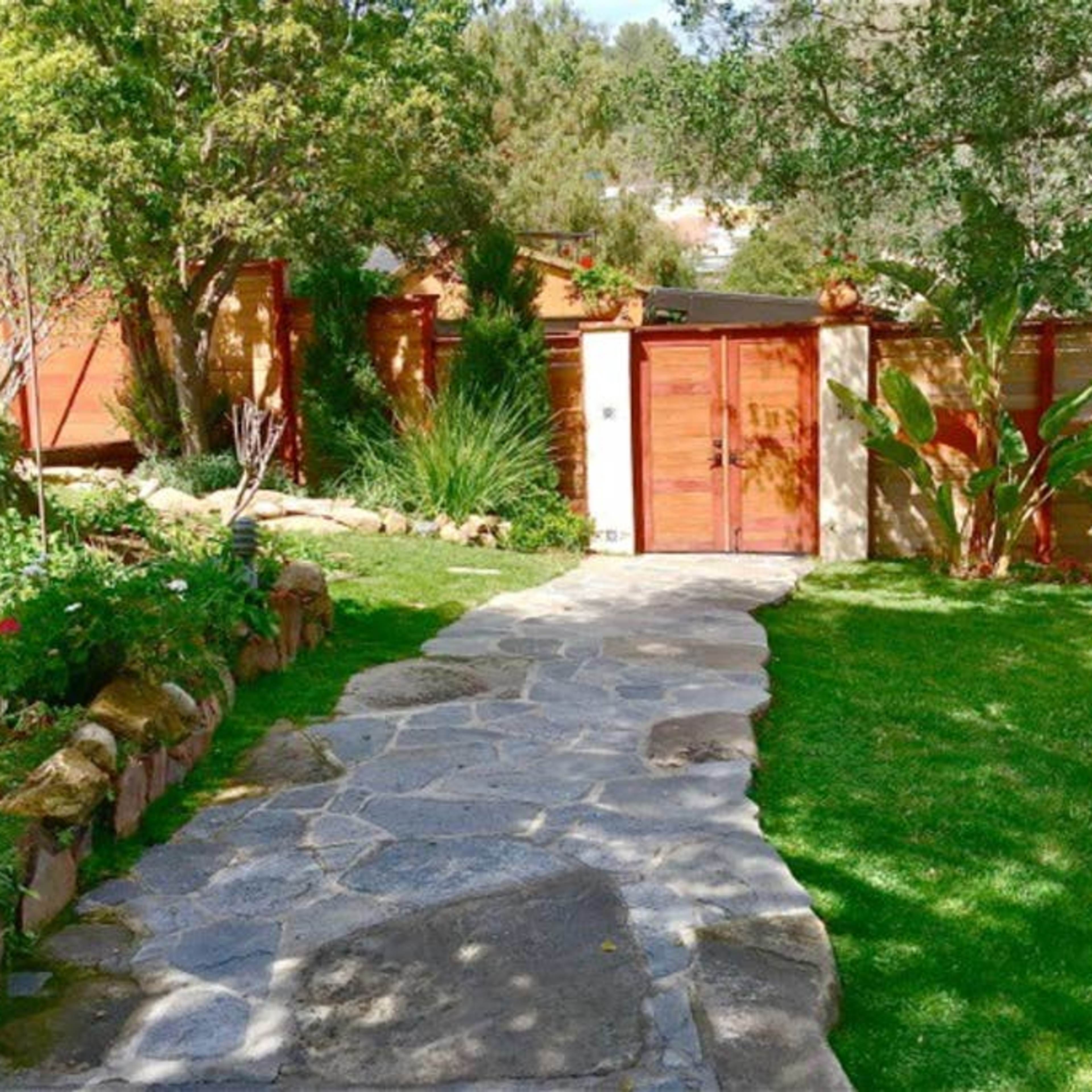 A stone pathway leads to a wooden gate surrounded by greenery and garden features.