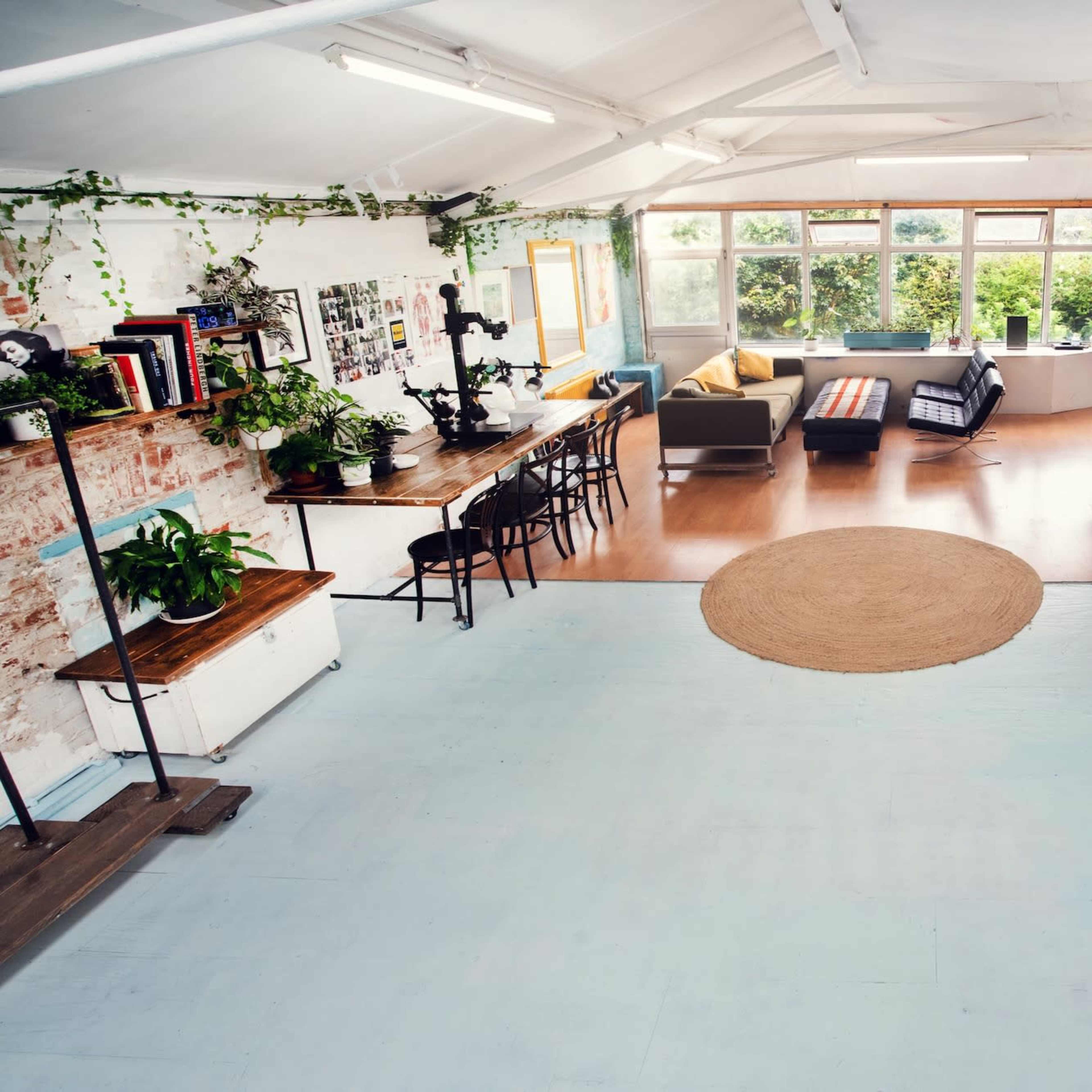 A spacious, well-lit studio with a mix of plants, a long dining table, seating areas, and large windows.
