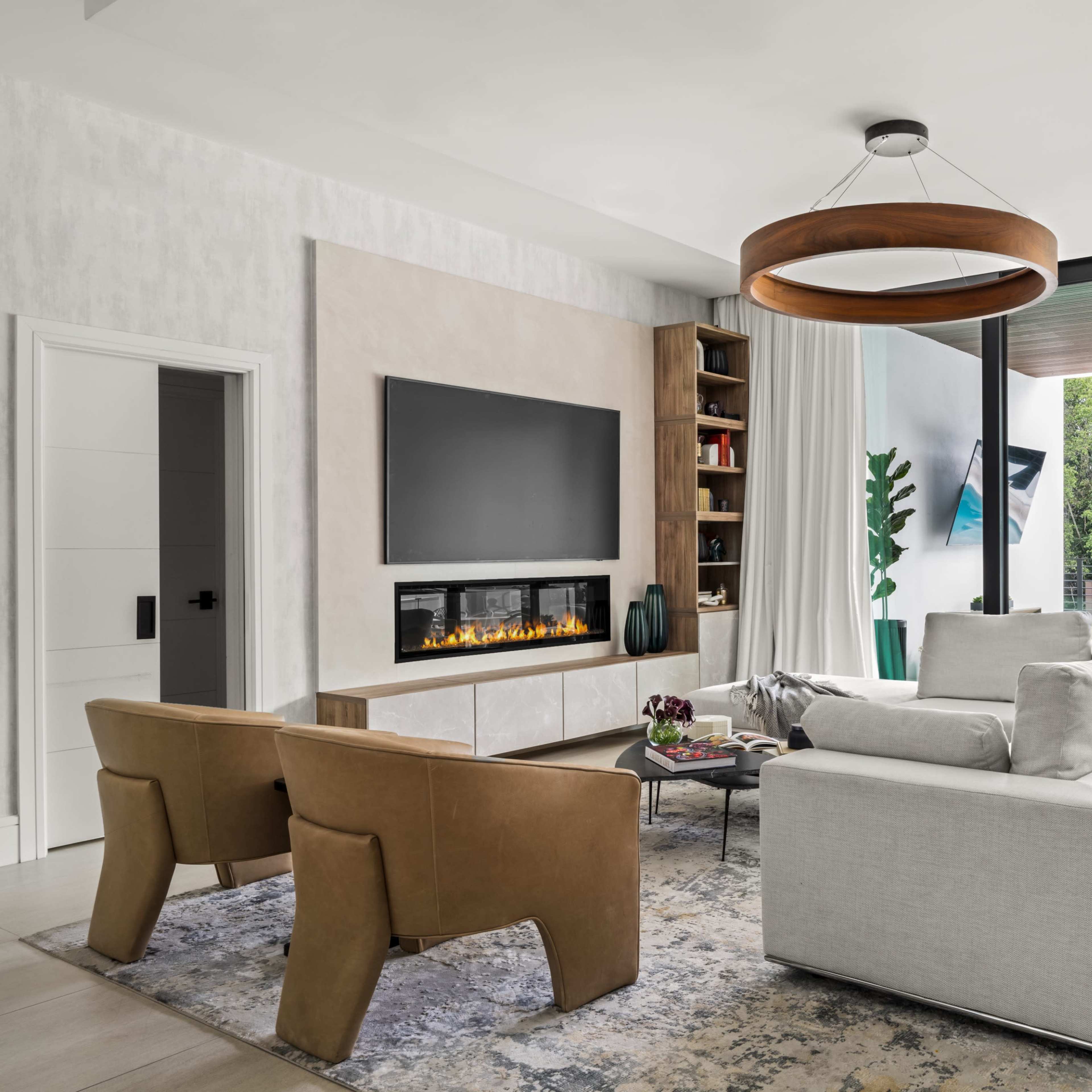 A modern living room features a sectional sofa, two armchairs, a wall-mounted television, and large glass windows overlooking a pool area.